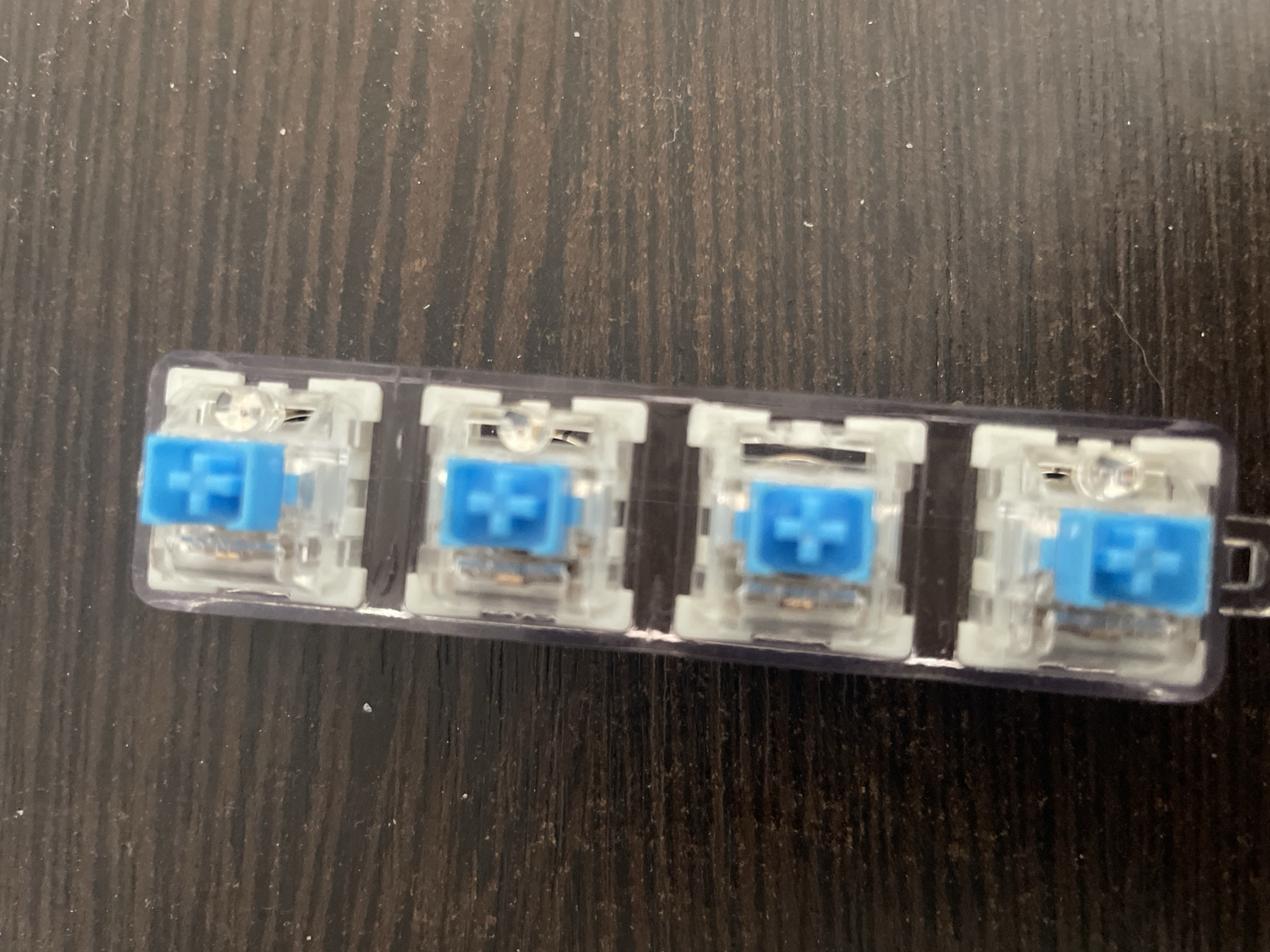The image shows a clear plastic strip holding four mechanical keyboard switches. Each switch has a translucent white housing and a bright blue cross-shaped stem, indicating the keycap mount. The switches are arranged in a row on a dark, textured surface, likely a wooden desk. The lighting appears to be artificial and overhead, casting subtle shadows. There is no visible text in the image. The overall scene is a close-up, static shot of electronic components, with no people or discernible environmental cues to suggest a specific time of day, weather, or location beyond the general context of a desk setup. The focus is solely on the keyboard switches, presented in a neat and organized manner within their packaging.