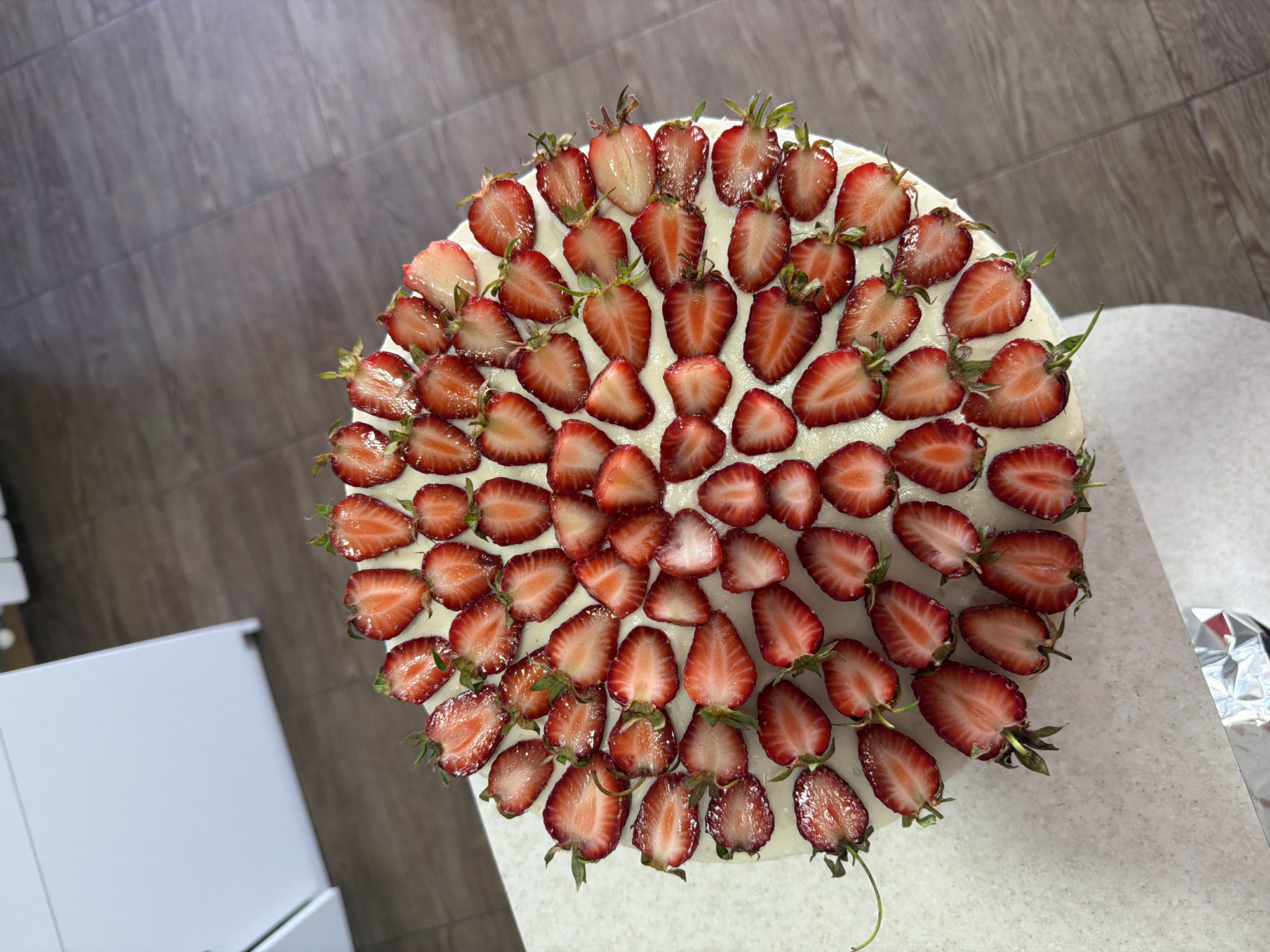 A top-down, medium shot reveals a strawberry-topped cake on a white surface, likely a table or countertop. The cake is circular and covered with a layer of white frosting or cream. Halved strawberries are meticulously arranged on top, forming concentric circles, with their cut sides facing upward to showcase the vibrant red flesh and tiny seeds. Some strawberries still have their green stems attached. The surrounding environment is modern, with the surface partially visible showing a pale, speckled pattern. In the background, light gray flooring is visible. A white cabinet or appliance is seen partially on the lower left. Based on general context clues, the location could be a kitchen or dining area. Location is unavailable without more information.