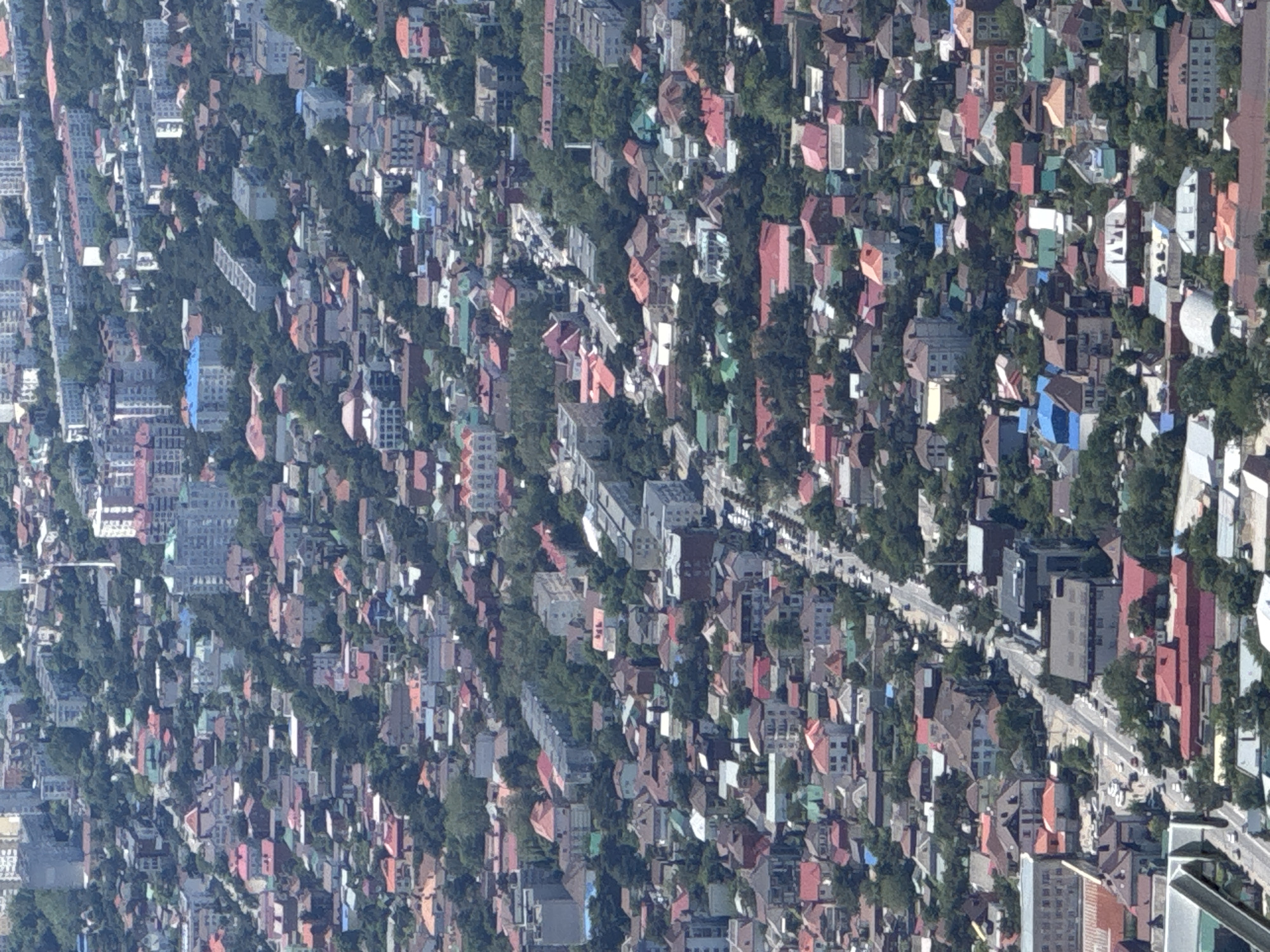 This image presents a rotated, high-angle aerial view of a densely populated urban area, consistent with the location context of Gelendzhik, Russia. The scene captures a vast expanse of residential and possibly commercial buildings interspersed with abundant greenery.
Numerous structures of varying sizes and architectural styles dominate the landscape. Many buildings feature distinctive pitched roofs in an array of colors, including terracotta red, orange, brown, grey, and occasional accents of blue or green. Taller, more modern apartment blocks with flat roofs are also visible, particularly towards the left in the current orientation of the image. Lush green trees and dense foliage are heavily integrated into the urban fabric, creating a patchwork of development and natural elements throughout the city.
Several roads and streets can be observed winding their way through the buildings and trees, with small vehicles discernible on them, indicating ongoing daily traffic. The image depicts a clear and bright daytime scene, with visible shadows suggesting the sun is high, likely in the mid-morning or early afternoon. The weather appears sunny and cloudless. No specific event or activity is taking place beyond the general rhythm of city life. There are no identifiable people or visible text within the frame.