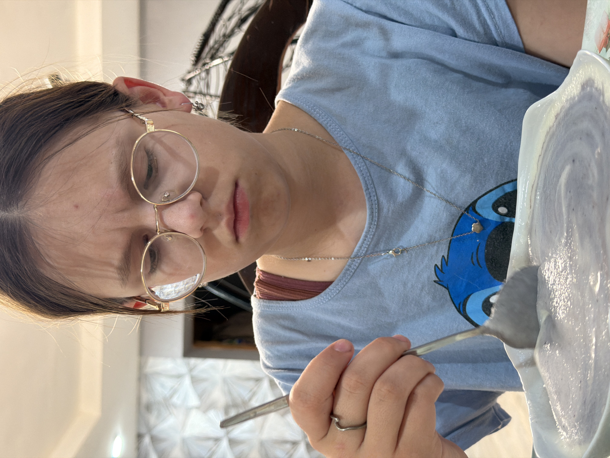 A young person wearing round eyeglasses and a blue t-shirt is holding a spoon near a bowl containing a white substance, possibly food. The person also has a silver necklace and a ring on their finger. The location is not discernible as Chaikovsky, Russia, from the visual information provided.