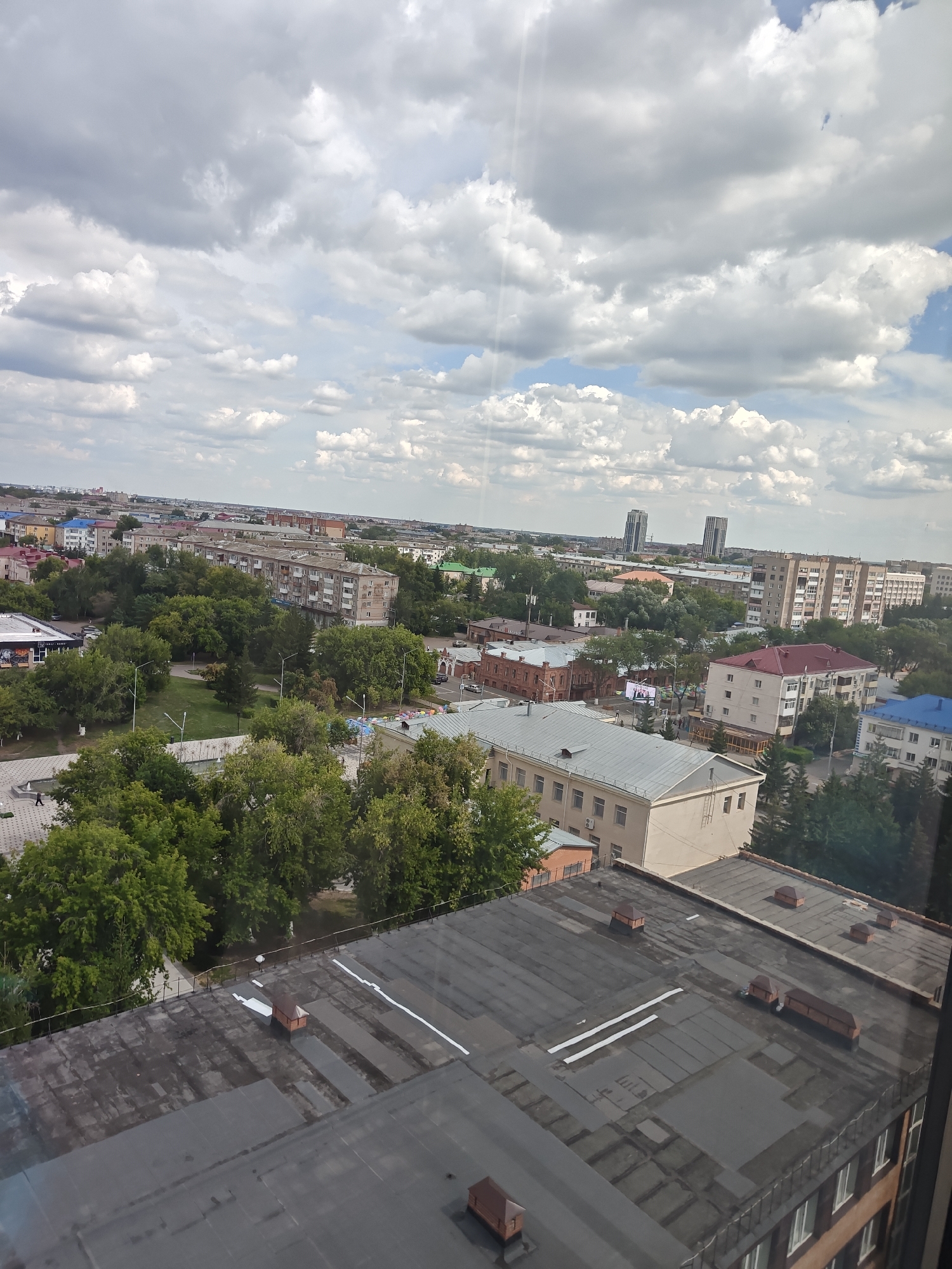 The image presents an aerial view of a city, likely Petropavl, Kazakhstan, under a partially cloudy sky. The foreground is dominated by the flat, dark rooftops of several multi-story buildings. These rooftops are textured with visible seams and some appear to have maintenance access points or ventilation structures. One rooftop in the mid-foreground features a small, reddish-brown wooden structure.
Further into the scene, a city landscape unfolds. Green trees are interspersed among various buildings, ranging in color and architectural style, including apartments with colorful roofs and other structures with more muted facades. A park or green space with manicured lawns and pathways is visible, with at least one person walking in the distance. Streetlights are present along the park's pathways. Two tall, modern skyscrapers stand out in the background, piercing the skyline. The overall impression is of an urban environment with a mix of older and newer architecture, integrated with significant green spaces. The lighting suggests it is daytime, possibly late morning or early afternoon, given the quality of light and the shadows.