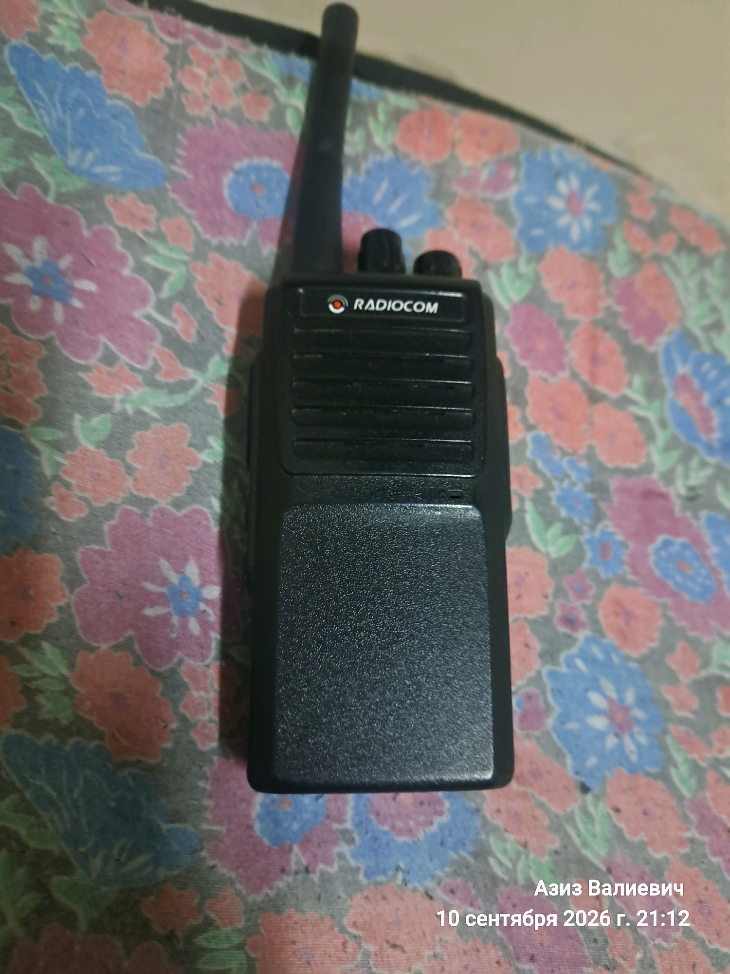 The image shows a black walkie-talkie with the brand "RADIOCOM" visible on its body, resting on a fabric with a floral pattern in shades of pink, blue, and green. The walkie-talkie is positioned vertically on the fabric, with its antenna extending upwards. In the lower right corner of the image, text in Cyrillic reads "Азиз Валиевич 10 сентября 2026 г. 21:12", which translates to "Aziz Valievich September 10, 2026 21:12". This text appears to be a timestamp and a name, possibly associated with the photograph or the object depicted. There is no discernible urban environment or specific location within Tashkent, Uzbekistan, visible in the image.
