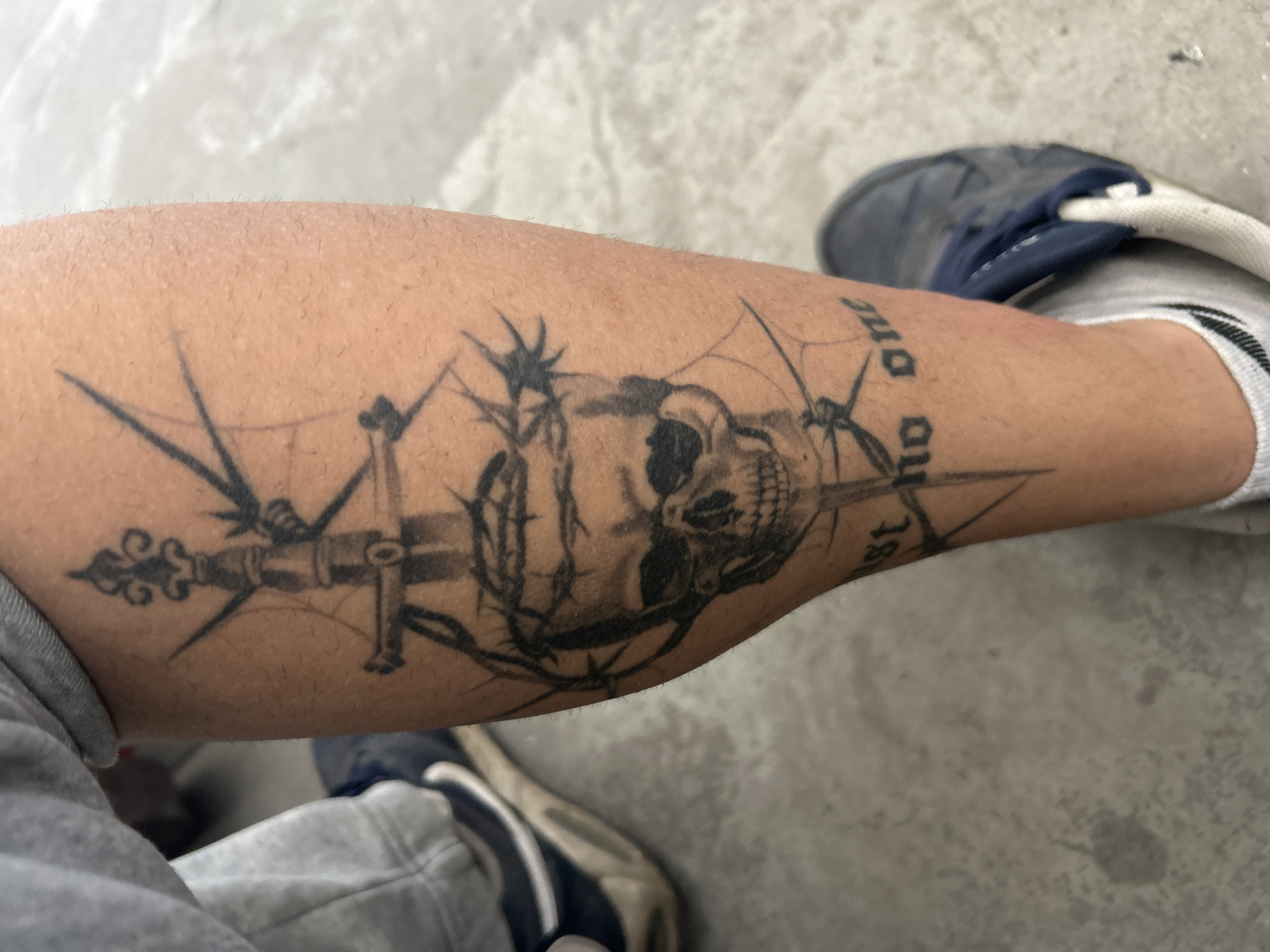 The image shows a person's lower leg with a tattoo. The tattoo features a skull with a sword or blade piercing through it. There is also what appears to be a crown of thorns surrounding the skull. The words "trust no one" are also incorporated into the design. The person is wearing a grey tracksuit and a blue and white shoe. The background is a grey concrete floor. The location is Unknown, Unknown.