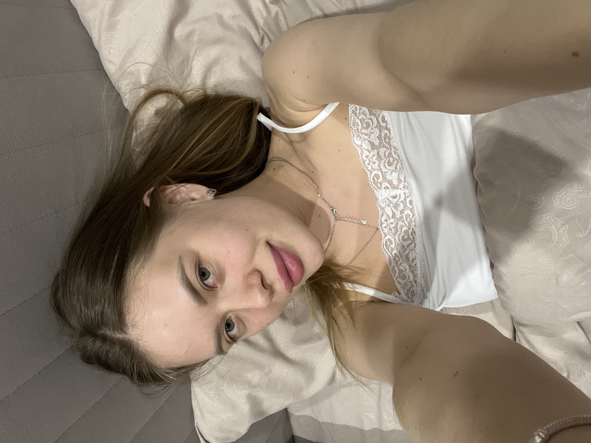 A young woman is captured in a close-up selfie, lying down, likely on a bed or sofa, in an indoor setting in Chaikovsky, Russia. She has long, light brown hair, which is casually spread around her head. She is wearing a white camisole with delicate lace trim along the neckline. A silver necklace with a small drop-shaped pendant is visible around her neck, and a silver ring adorns her right pinky finger, along with a bracelet on her wrist.
Her head is slightly tilted, and she gazes directly at the camera with a relaxed expression, featuring light green or hazel eyes and a faint, gentle smile with her lips slightly parted. The background includes light-colored bedding, consisting of a pillow and what appears to be a duvet or sheet with a subtle pattern. To her left, a gray, possibly padded, headboard or sofa backrest is visible. The lighting is soft and even, suggesting a comfortable, personal space. The overall scene conveys a casual and intimate moment of self-portraiture. No text is visible in the image.
