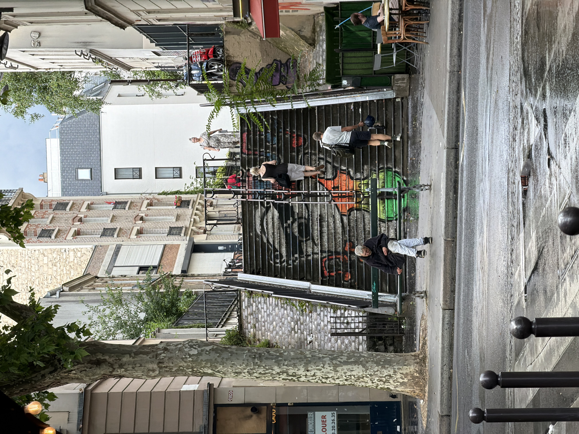 The image depicts a street scene in Paris, France. In the foreground, a wet asphalt street is visible with dark bollards lining the sidewalk. Further back, a man sits on a park bench in front of a set of graffiti-covered stairs. Several people are ascending the stairs. Behind the stairs, there are buildings and trees. Two scooters are parked near the top of the stairs. In the background, a person is seated at a table outside a cafe. The overall atmosphere suggests a cloudy day, with evidence of recent rain.