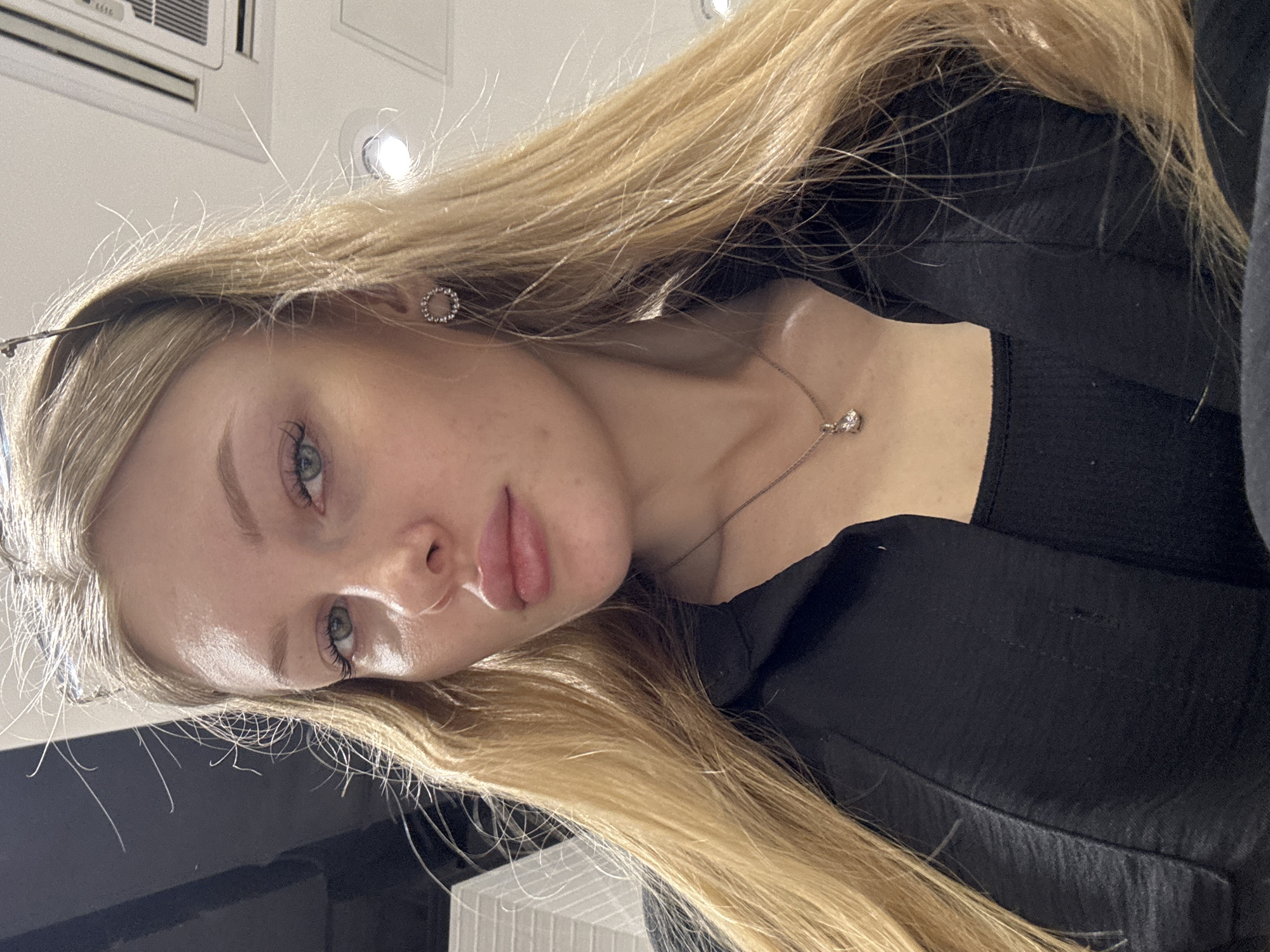 A young, fair-skinned woman with light blonde hair is centrally positioned in the image. Her head is tilted to the right. She has blue eyes. She is wearing a silver necklace with a pendant and a small silver earring in her left ear. She is dressed in a black top or blouse. The background is indistinct, but suggests an indoor setting, likely a building or office, potentially in Perm, Russia, based on likely metadata or image content. An air conditioning unit and ceiling lights are visible in the upper part of the frame. The image appears to be taken at a close range.