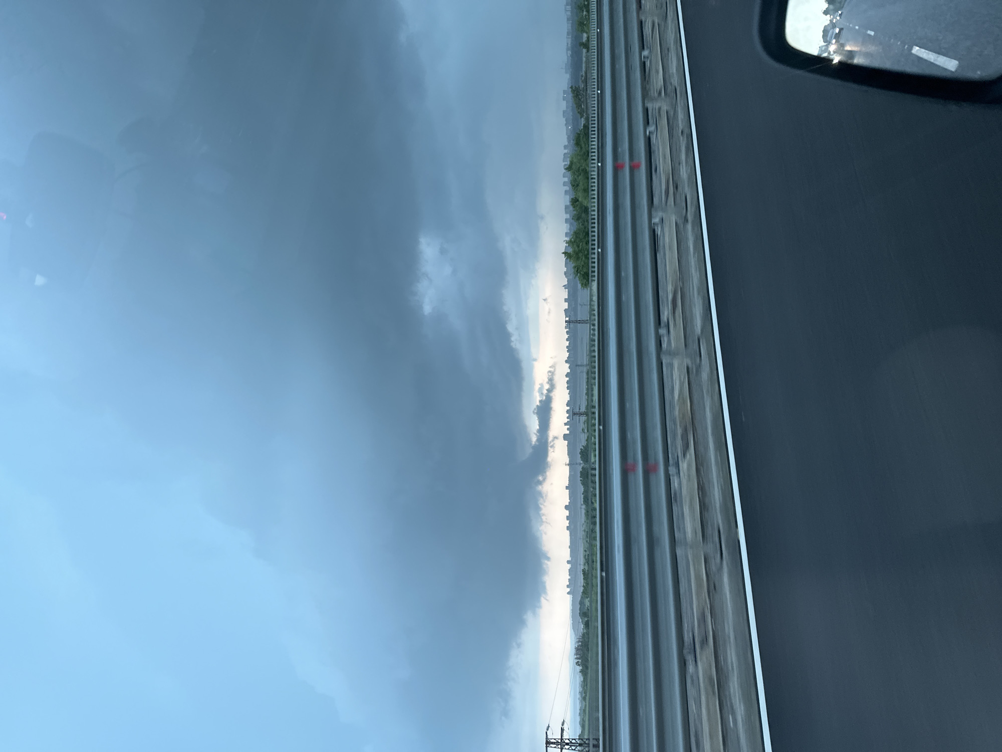 The image, taken from inside a vehicle, captures a landscape scene under heavily overcast skies, suggesting stormy weather. The view appears to be from the passenger side, looking forward and to the left.
The upper portion of the frame is dominated by a vast, cloudy sky, ranging from light blue to dark, ominous grey, particularly dense towards the center and horizon. A brighter, lighter strip of sky is visible just above the distant landscape, contrasting with the darker cloud mass above.
In the lower half of the image, a multi-lane highway runs horizontally, parallel to the horizon line. The road surface appears wet, indicating recent or ongoing rain. Guardrails or barriers line the right side of the highway, showing vertical slats and some horizontal elements. Several small red markers or reflectors are visible on the road surface or barriers.
Beyond the highway, a strip of green trees and foliage is visible, separating the road from a faint, distant cityscape or skyline of buildings. To the far left, a metal utility pole or tower is partially visible against the cloudy background.
On the far right edge of the image, the vehicle's side-view mirror is partially visible, reflecting the highway behind, with blurred lights from other vehicles suggesting traffic. The left side of the image shows a blurry reflection from inside the vehicle's cabin, likely on the windshield.
The overall impression is of driving on a highway during a gloomy, rainy day, with dark storm clouds gathering over a distant urban area.