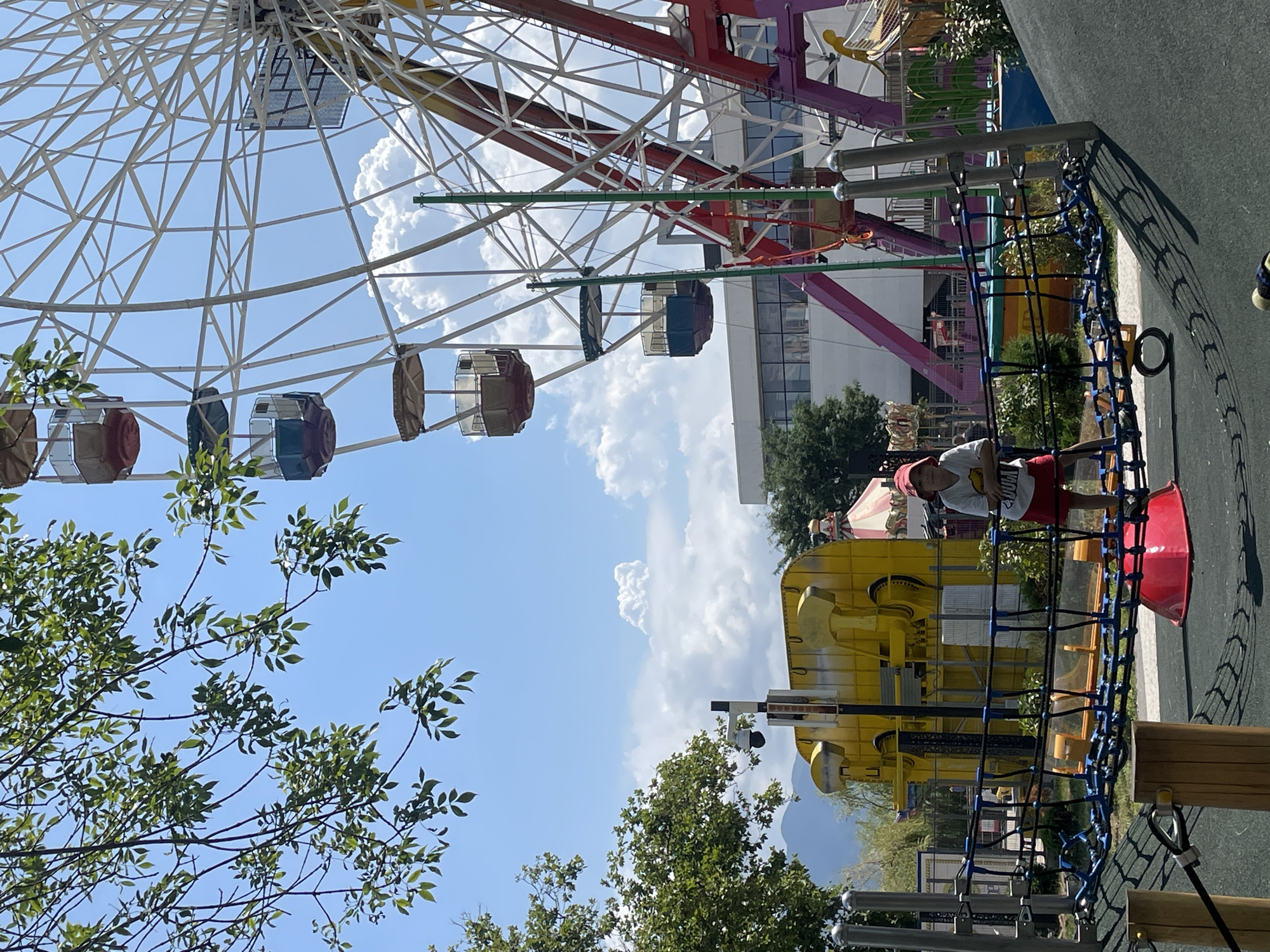 This image captures a vibrant amusement park scene on a bright, sunny day, likely located in Novorossiysk, Russia, given the context. In the foreground, a young boy, wearing a red hat and red shorts with a white t-shirt, stands on a blue and black rope bridge. The bridge is part of a children's playground area with a dark, rubberized surface. Behind the playground, a large, colorful Ferris wheel dominates the background, with several gondolas visible against a partly cloudy sky. To the left of the Ferris wheel, a large, yellow amusement park ride or structure is visible, along with parts of other attractions including a carousel. Green trees and foliage frame the scene, and in the distance, mountains can be seen. The overall atmosphere is one of leisure and outdoor fun.
