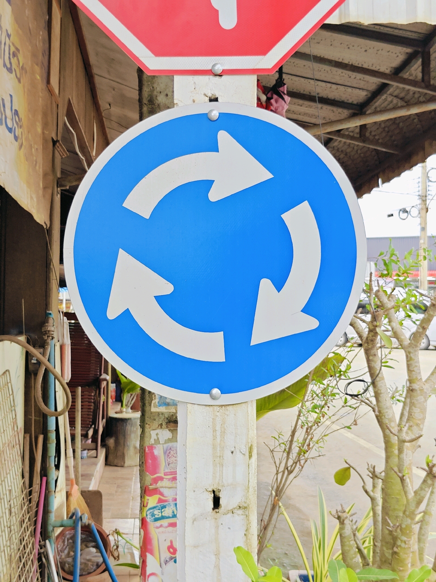 A blue, circular traffic sign with three white arrows pointing in a clockwise direction is mounted on a white post in Tambon Mu Si, Thailand. The sign is positioned in front of a building with a wooden facade and a corrugated metal roof. To the left of the sign, various objects are leaning against the building, including a rake, a broom, and some pipes. To the right of the sign, a tree with sparse foliage and some smaller plants are visible. In the background, a road and several parked vehicles can be seen. The overall scene appears to be a street in a somewhat rural or developing area.