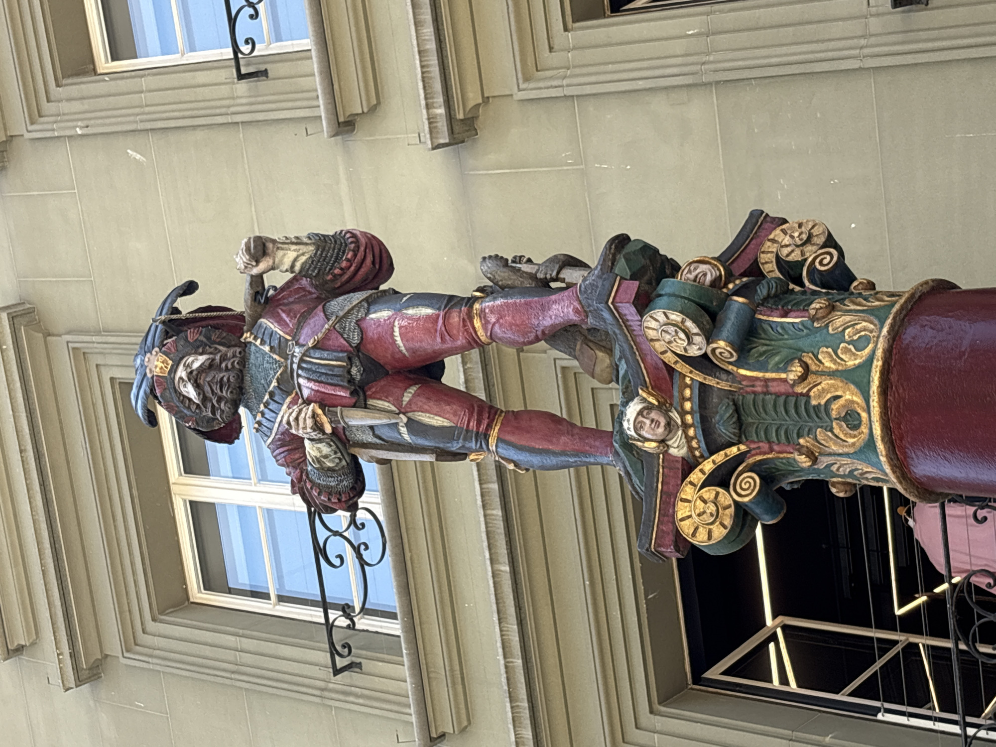 The image shows a detailed, colorful wooden sculpture of a figure in ornate attire, possibly a historical or theatrical costume, suspended from a building. The figure is depicted in a dynamic pose, with its legs extended and one arm bent at the elbow, holding something indistinct. The sculpture is mounted on a richly decorated column featuring intricate carvings of faces, leaves, and scrollwork, painted in green, gold, and red. A large, dark red cylindrical element extends from the column to the right of the frame.
The sculpture is positioned in front of a light-colored stone building with a large window featuring a grid of panes. The window reflects a bright blue sky, suggesting daylight. The building architecture, with its classical molding and stone blocks, indicates an urban setting with historical significance. A black wrought-iron decorative element is attached to the building, partially overlapping the sculpture. The overall impression is of a decorative architectural element or a street art installation. The lighting suggests it is daytime, and the clear reflection in the window implies fair weather. There is no visible text in the image. The location context suggests this is in Bern, Switzerland.