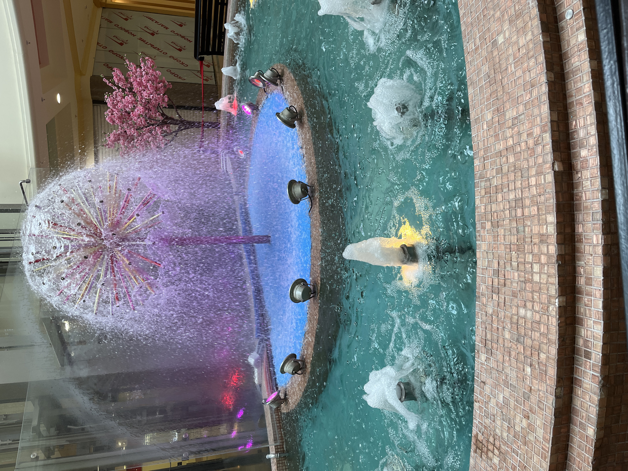 The image shows a fountain in an indoor location. The fountain has a central, spherical water feature with multiple water jets and is lit with pink and purple lights. In the background, there is a faux pink tree. The fountain is surrounded by a pool of turquoise water. The pool's edge is lined with lights. The wall next to the pool is tiled with brown and beige squares. The location is in an unknown city.