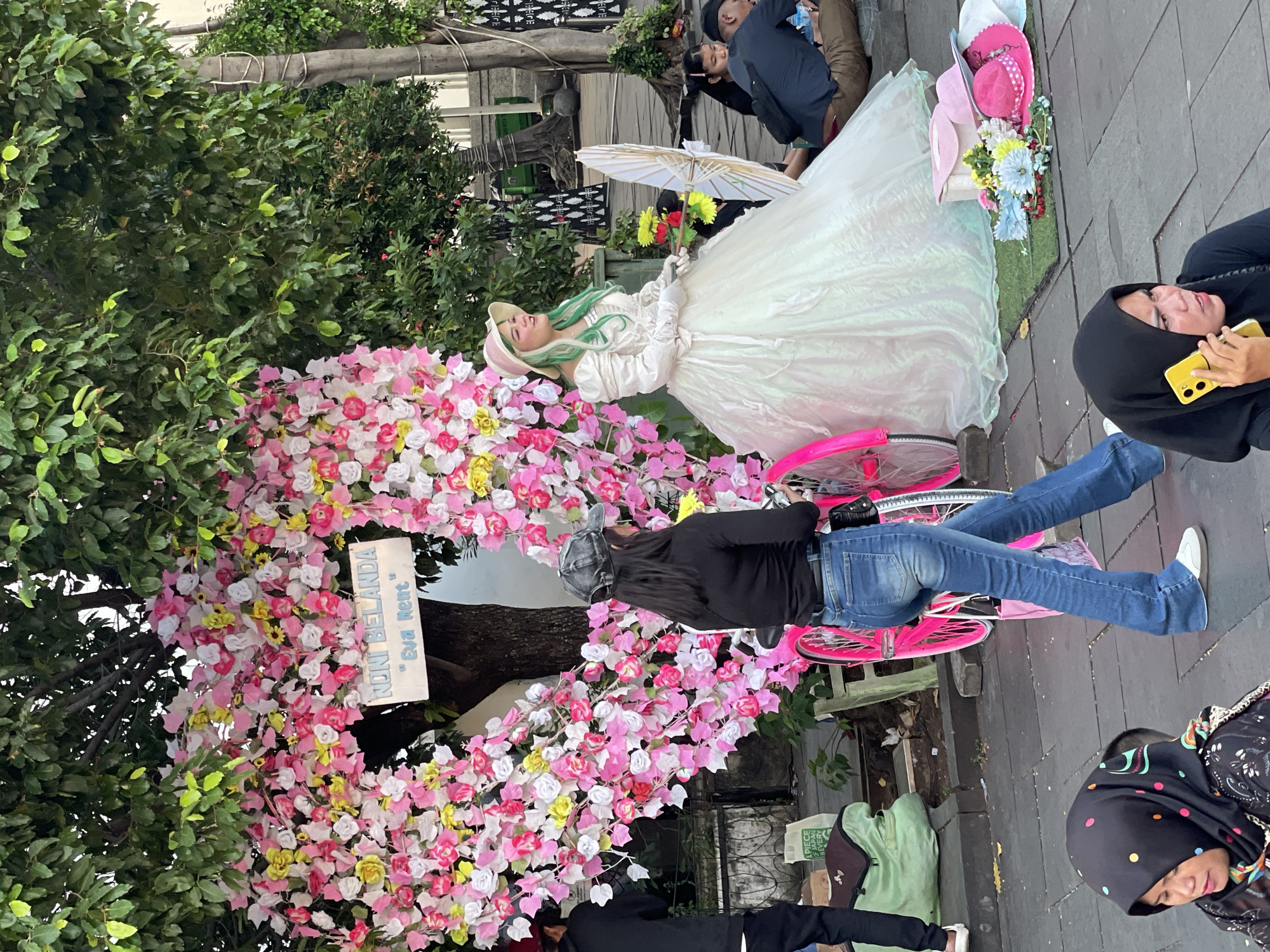 In a bustling pedestrian area in Kota Tua, West Jakarta, Indonesia, a young woman dressed in a voluminous, iridescent white gown and a pink hat with green-dyed hair poses for photos. She is seated on the ground next to a large, heart-shaped floral arrangement made of pink, white, and yellow flowers, intertwined with the branches of a mature tree. A bright pink bicycle with a white umbrella is positioned behind her.
Several other individuals are present in the scene. To the left, a person wearing a black hoodie and black pants stands, holding a camera. To the right, a woman in blue jeans and a black long-sleeved shirt, wearing a grey baseball cap, walks past the floral heart, looking towards the posed woman. In the background, a few people are seated on the ground, and a person in a black hijab is seen talking on a yellow smartphone. The ground is paved with square tiles, and the area is shaded by the foliage of the trees.