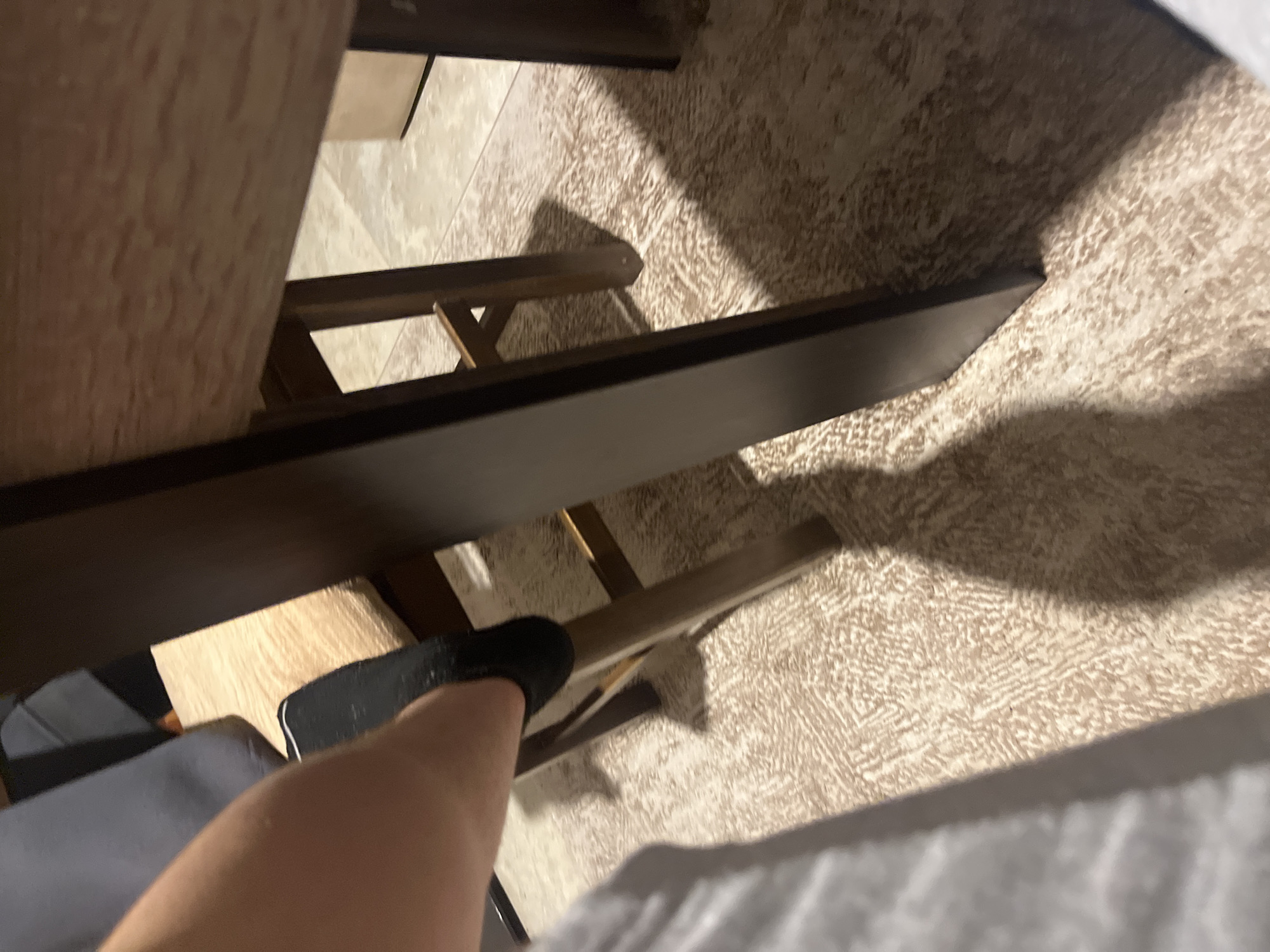 The image depicts an indoor scene in Blagoevgrad, Bulgaria, observed from a low angle. A bare human forearm is visible in the upper-left quadrant. Adjacent to the arm, a section of grey fabric is present. In the central area, a foot, covered by a black sock with a white stripe near the ankle, is positioned against a dark brown wooden chair. The chair features a light beige, textured seat cushion and dark brown wooden legs and horizontal rungs. To the right, a substantial, dark brown leg of a table or desk is vertically oriented. The floor in the foreground is covered by a patterned light beige and brown carpet, which transitions to a light-colored tiled floor further into the background. Distinct, elongated shadows from the furniture are cast upon the carpet, indicating a singular light source.