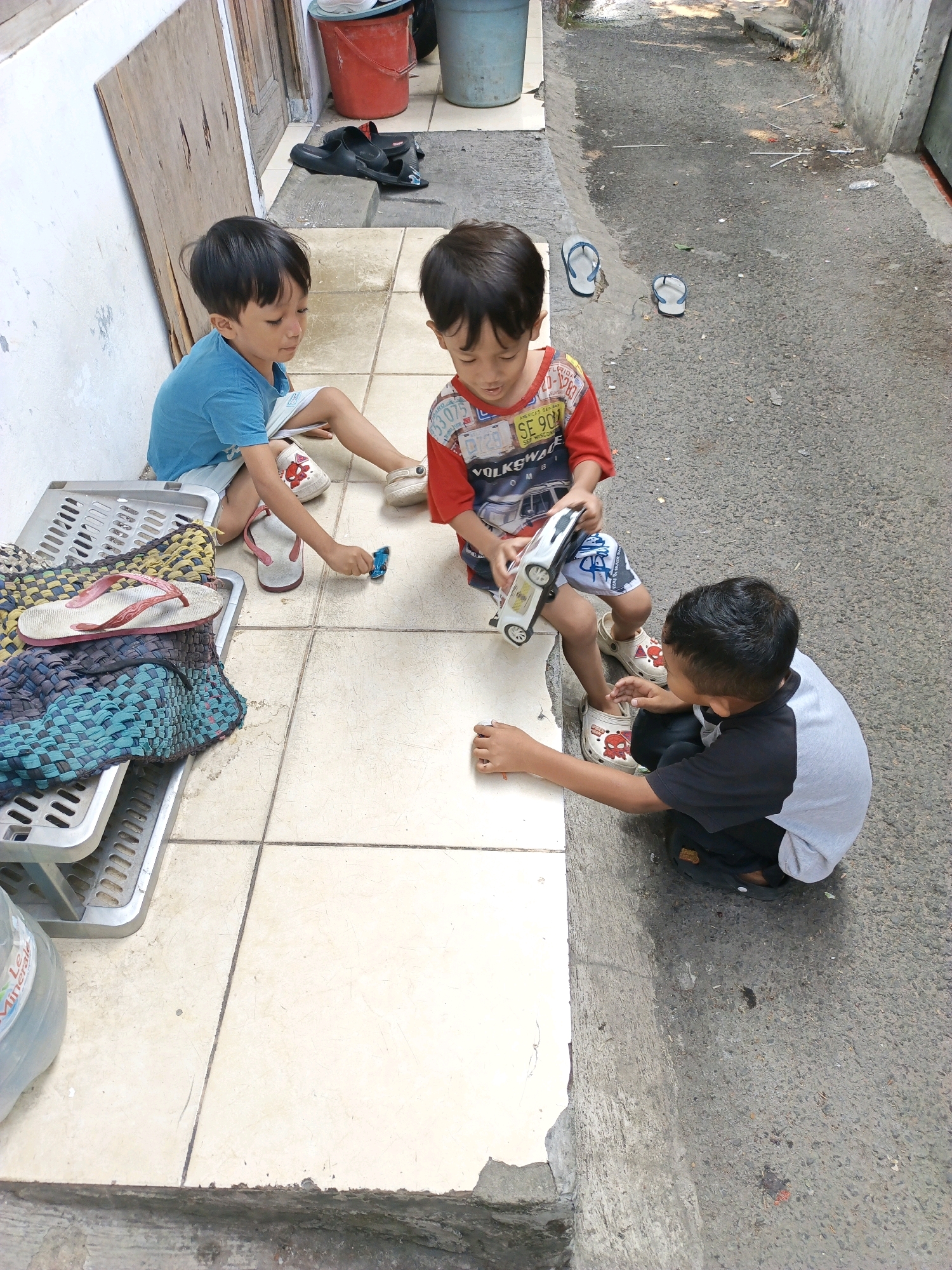 Three young boys are playing with toys on a tiled pathway next to an asphalt road. The boy on the left, wearing a blue t-shirt and shorts, is sitting and holding a small blue toy. The boy in the middle, wearing a red and white patterned t-shirt and shorts, is sitting and holding a white toy car. The boy on the right, wearing a black and grey t-shirt and black shorts, is squatting and reaching out to the toy car. In the background, there are several pairs of flip-flops, a red bucket, a blue bucket, and a woven mat on a plastic rack. The scene appears to be an outdoor alleyway in a residential area of South Jakarta, Indonesia.