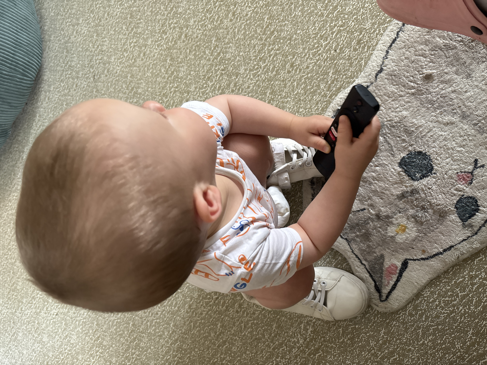A baby is squatting on a carpeted floor in what appears to be a residential setting. The baby is wearing a white onesie with an orange and blue graphic pattern, and white sneakers. The baby is holding a black remote control in their hands and is looking down at it. A small rug with a cat design is visible on the floor in front of the baby. There is no visible indication of the location being in Naberezhnye Chelny, Russia. The environment is a well-lit indoor space with a textured carpet.