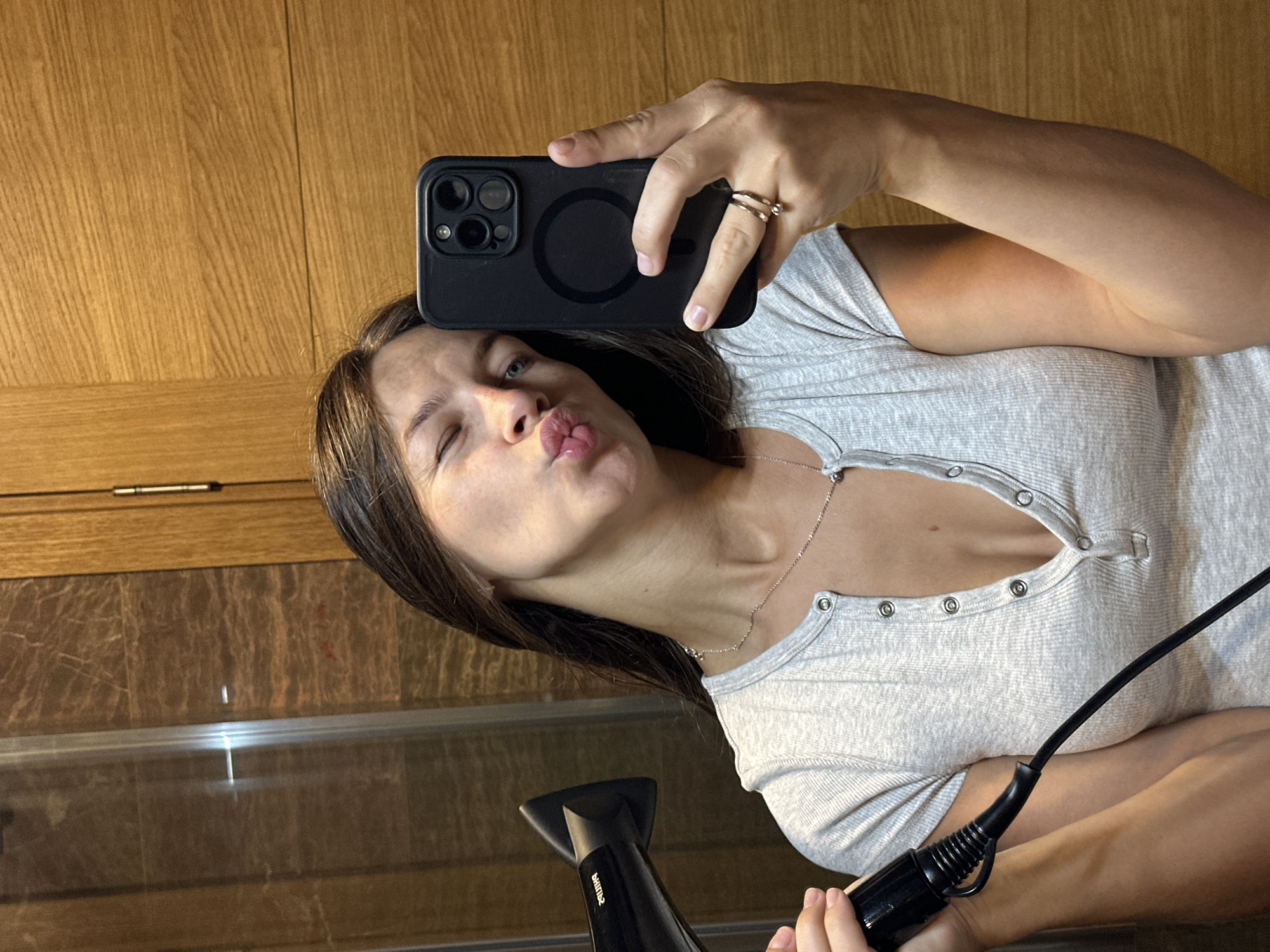 A young woman is taking a selfie in a bathroom. She is wearing a grey Henley shirt and has her hair pulled back. She is holding a black hairdryer in her left hand and a black smartphone in her right hand, which she is using to take the picture. Her lips are puckered, and she is winking at the camera. The background appears to be a modern bathroom with marble tiles and wooden accents. The scene suggests a private, domestic setting, likely in a home or hotel. The location of Donji Pobori, Montenegro is not discernible from the visual information presented in the image.