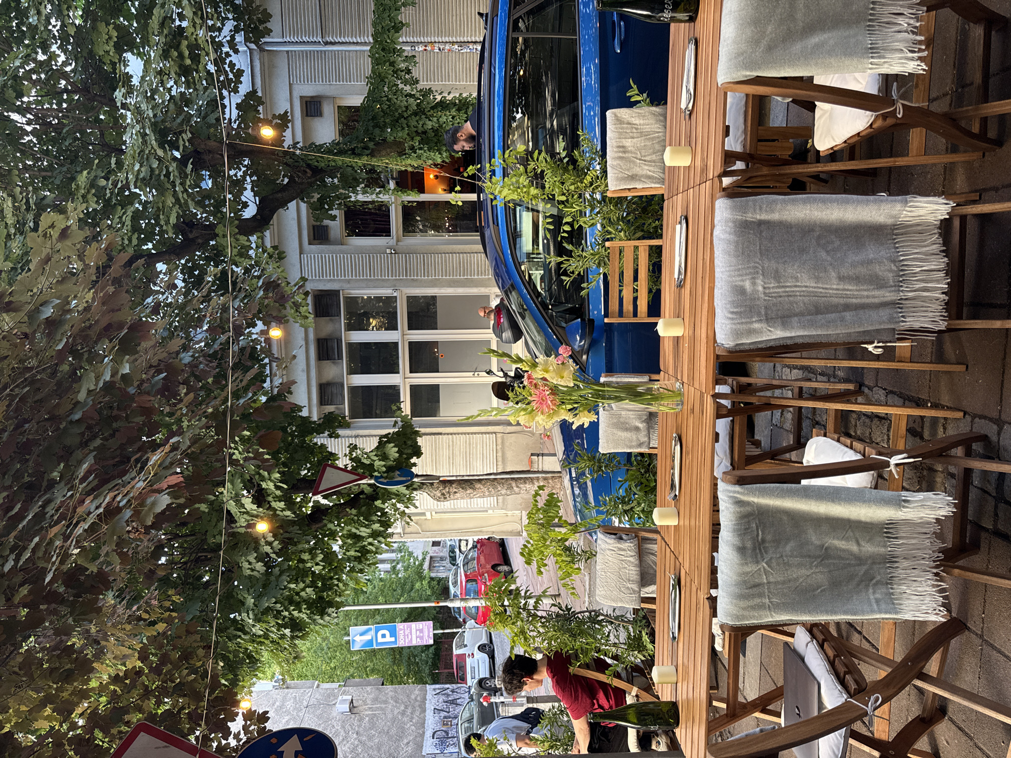The image captures an outdoor seating area of a restaurant on a street in Belgrade, Serbia, likely in the evening. A long, wooden table is set for dining with white placemats, cutlery, and candles. Two chairs draped with light grey blankets with white fringe are visible in the foreground. A person wearing a red shirt is seated at the far end of the table, and a bottle of green liquid, possibly wine, stands on the table. Behind the table, a bright blue car is parked on the street, with a person leaning out of its sunroof. Trees with lush green foliage overhang the street, with string lights illuminating the scene. Buildings with windows and architectural details line the street. Traffic signs and parked cars are also visible in the background.