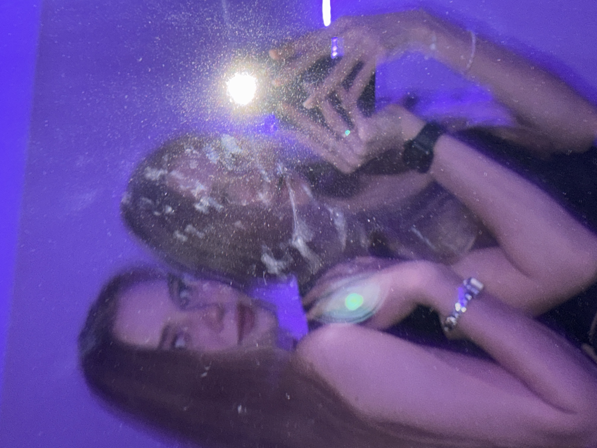 The image shows a mirror selfie taken by two young women. The woman on the left is facing the camera with long brown hair and red lipstick. She is wearing a black top. The woman on the right is holding a phone to take the picture, with her face partially obscured. She is also wearing a black top and a watch on her left wrist. Both women are wearing bracelets. The background is a blurred purple color. The image appears to be taken indoors, possibly in a bedroom or bathroom, as suggested by the presence of a mirror and the intimate nature of the selfie. There is no discernible information in the image that would allow for its localization in the city of Perm, Russia.