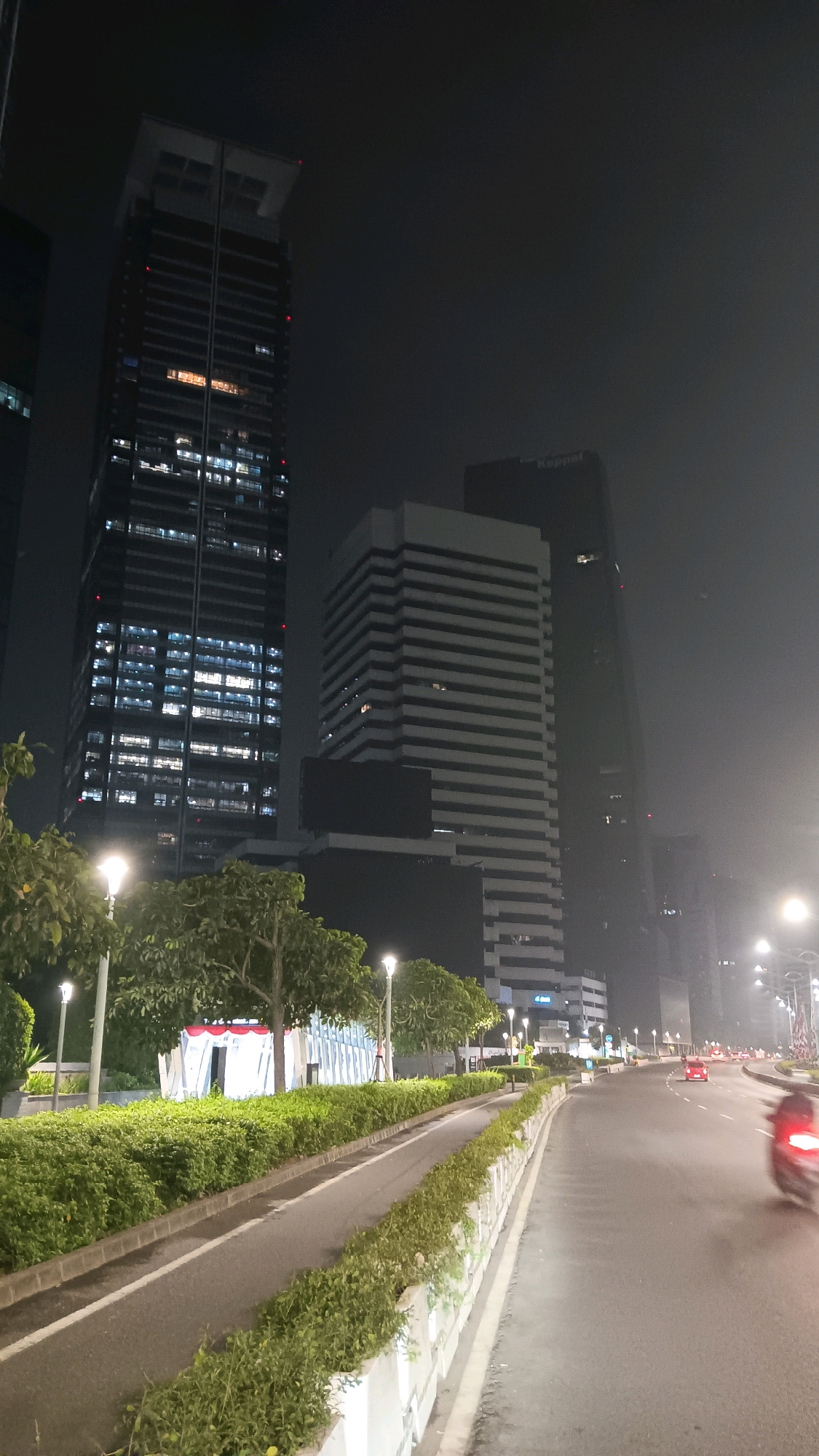 A nighttime, slightly hazy exterior shot captures a view of the city of South Jakarta, Indonesia. The foreground shows a paved road curving to the right, bordered by manicured green hedges and concrete barriers. A bicycling lane is visible on the left side of the road, also lined with hedges and streetlights. Several trees with lush green foliage are interspersed along the roadside. In the middle ground, a modern building with a white facade and dark horizontal stripes stands prominently, with a taller, darker skyscraper directly behind it. The skyscraper on the left has numerous illuminated windows, indicating it is occupied. A red car is visible in the distance on the road, and a blurred figure on a motorcycle is in the right foreground, suggesting motion. The overall impression is of an urban environment at night, with city lights and tall buildings illuminated against a dark sky.