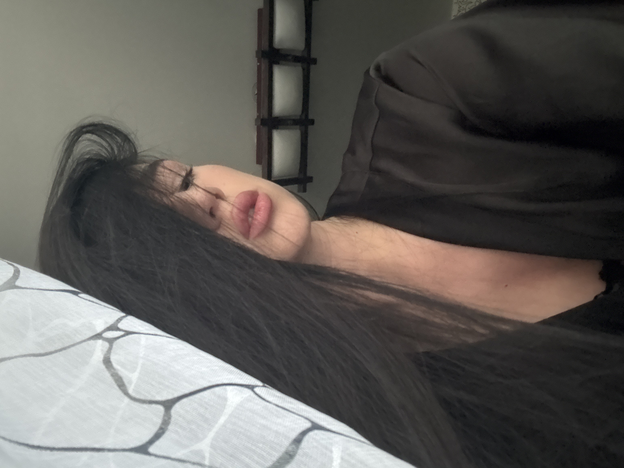 The image shows a close-up of a woman's face from the chest up, in a dark room. She has long, dark hair that partially obscures her face, and her eyes are closed. She is wearing a dark robe. A patterned sheer curtain is visible on the left side of the image. In the background, a light fixture with three rectangular lamps is mounted on the ceiling. The image does not provide any information that would allow for determining its location in Novosibirsk, Russia.