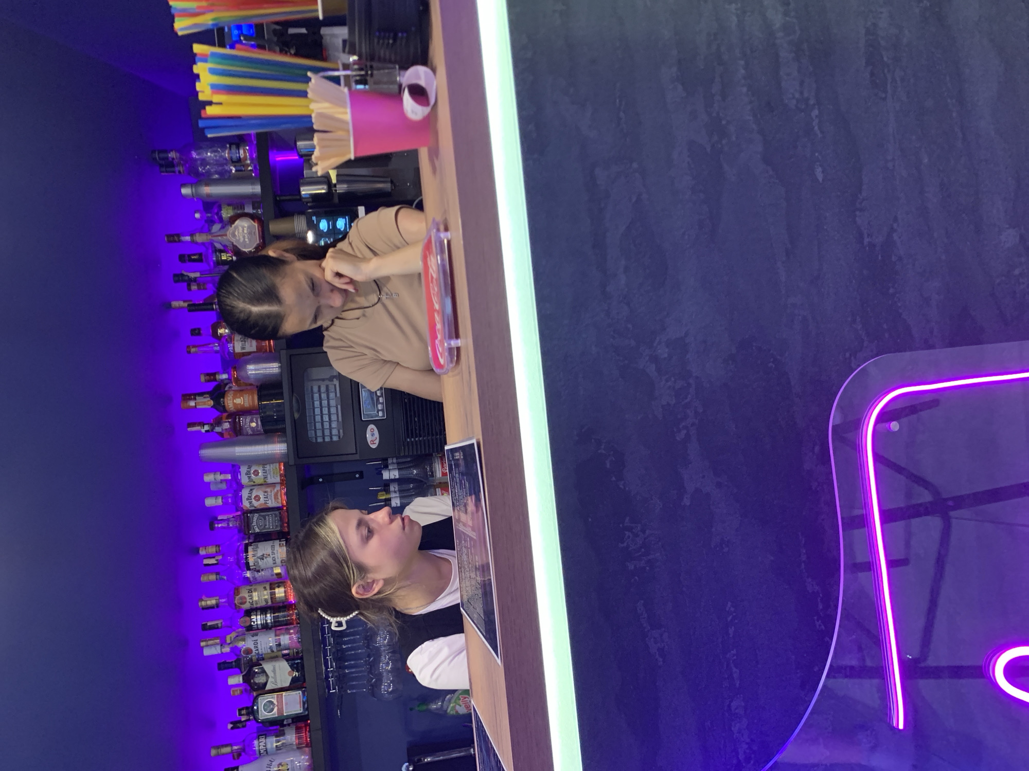 In the city of Kapyĺ, Belarus, this image captures an indoor scene likely within a bar or establishment with a bar area. The primary focus is on two young women, presumably staff members, standing behind a bar counter. The woman on the left, with blonde hair styled with a white clip, is wearing a white t-shirt and a dark vest. She is looking down at a menu or document on the counter, her expression suggesting concentration. To her right, a woman with dark hair is also looking down, resting her chin on her hand, appearing somewhat pensive or bored. She is wearing a light brown t-shirt.
The bar counter itself is made of wood, with a bright green neon light strip illuminating its edge. Behind the counter, shelves are lined with various bottles of alcohol, predominantly wine and spirits, illuminated by purple ambient lighting from above. Various bar accessories are visible, including glassware hanging upside down, a small black machine, and a bright pink cup filled with colorful drinking straws. On the counter, a red Coca-Cola branded item is placed on a transparent stand, and a roll of white tape is also visible. The background wall is painted a deep purple. The lower portion of the image shows a textured dark surface, possibly a wall or furniture, with a prominent purple neon sign in the foreground. The overall atmosphere suggests a dimly lit, modern bar environment.