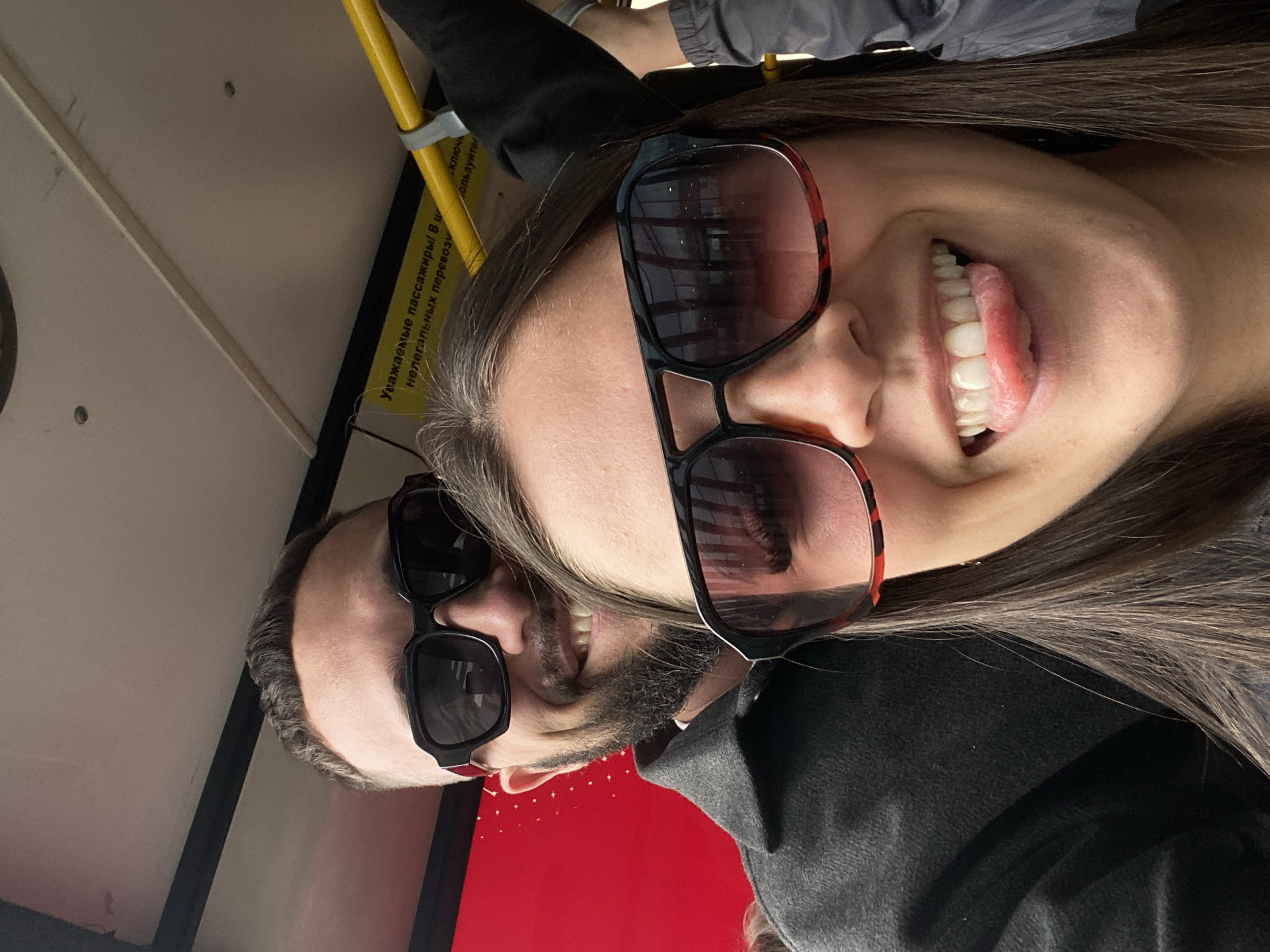 This is a selfie taken by two people, a man and a woman, on what appears to be a public transport vehicle, possibly a bus. The image is taken from a low angle, and the two individuals are positioned close together, with the woman on the right and the man on the left. Both are wearing sunglasses, and the woman is sticking her tongue out, suggesting a playful or humorous mood. The woman has long dark hair, and the man has short brown hair and a beard.
The background is somewhat blurry but shows interior elements of the vehicle, including a yellow handrail and some yellow signage with text in Russian. The overall lighting suggests it is daytime. The presence of the handrail and the yellow sign with text indicates they are likely in Russia. The text on the yellow sign reads: "Уважаемые пассажиры! В целях вашей безопасности, перед началом движения, во время движения и при выходе из транспортного средства, держитесь за поручни." This translates to: "Dear passengers! For your safety, before the start of movement, during movement and when exiting the vehicle, hold on to the handrails."
The scene is casual and intimate, with the subjects appearing relaxed and enjoying themselves. The focus is on their faces and expressions, conveying a sense of fun and connection.