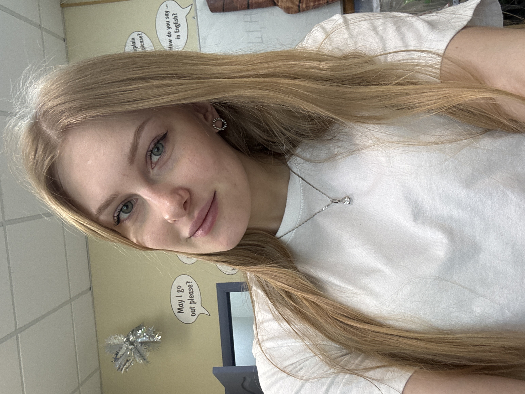 A close-up portrait shows a young woman with long, light-brown hair and light eyes. She wears a white shirt and a silver necklace with a diamond pendant. She has round earrings on her left ear. Behind her, a wall is visible with educational posters in English, with phrases such as "How do you say… in English?" and "May I go out please?". A silver decorative snowflake is also visible on the wall. A mirror and part of a desk are visible in the background. There is no evidence to confirm the location is in the city of Perm, Russia.