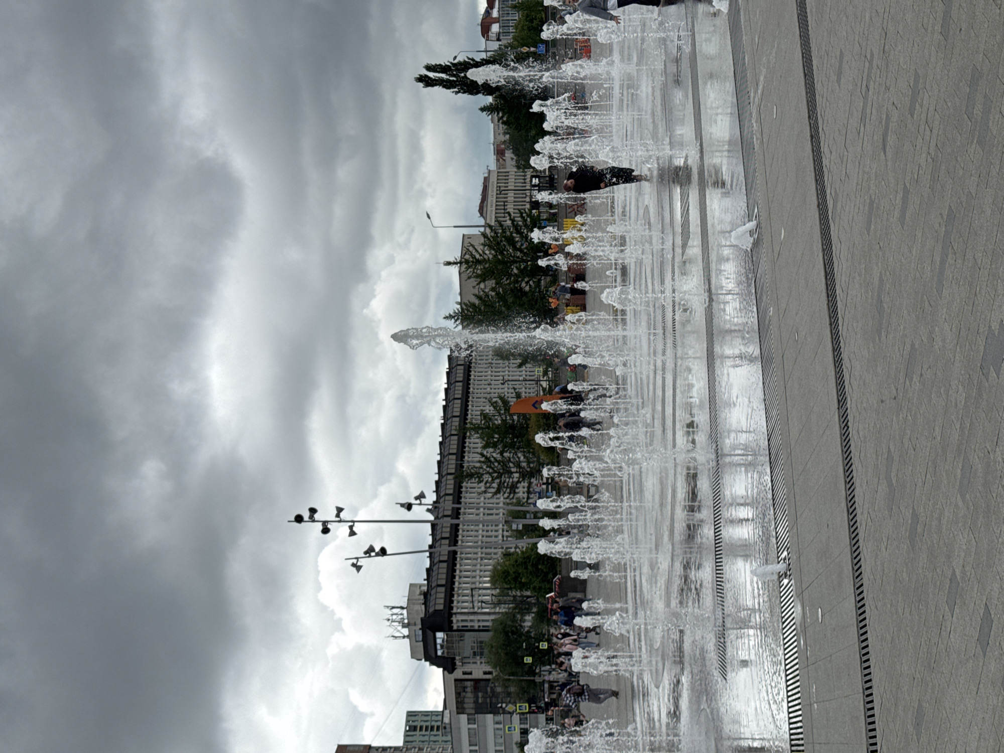 The image depicts a wide-angle outdoor scene in the city of Perm, Russia, under an overcast sky filled with dense grey clouds. The foreground consists of light grey paving stones, some arranged in a patterned brickwork, with multiple parallel linear drainage grates running horizontally across the lower third of the frame.
A large, ground-level interactive fountain occupies the mid-ground. Numerous distinct jets of clear water of varying heights erupt directly from the paved surface, creating a dynamic water display. The water forms a shallow pool on the fountain surface, reflecting the grey sky and surrounding structures. Several individuals are present within the fountain area; one person in a dark jacket is visible mid-right, walking through the water, while another in a light grey top is partially visible on the far right edge. Other individuals are scattered around the periphery of the fountain on dry pavement. They are casually dressed and appear to be observing or passing through the area.
In the mid-background, a prominent multi-story building with a dark, flat roof and numerous rectangular windows is visible. Antenna structures are mounted on its roof. To its left, other buildings of varying architectural styles are discernible, including a taller, more modern structure. Several dark green coniferous trees are interspersed between the fountain and the buildings. Multiple tall, multi-headed lamp posts, featuring several conical fixtures radiating from a central vertical pole, are positioned in the mid-ground, extending above the treeline. Additional standard streetlights are also present. The scene captures a public urban space with human activity centered around the fountain, despite the muted lighting from the heavily clouded sky.