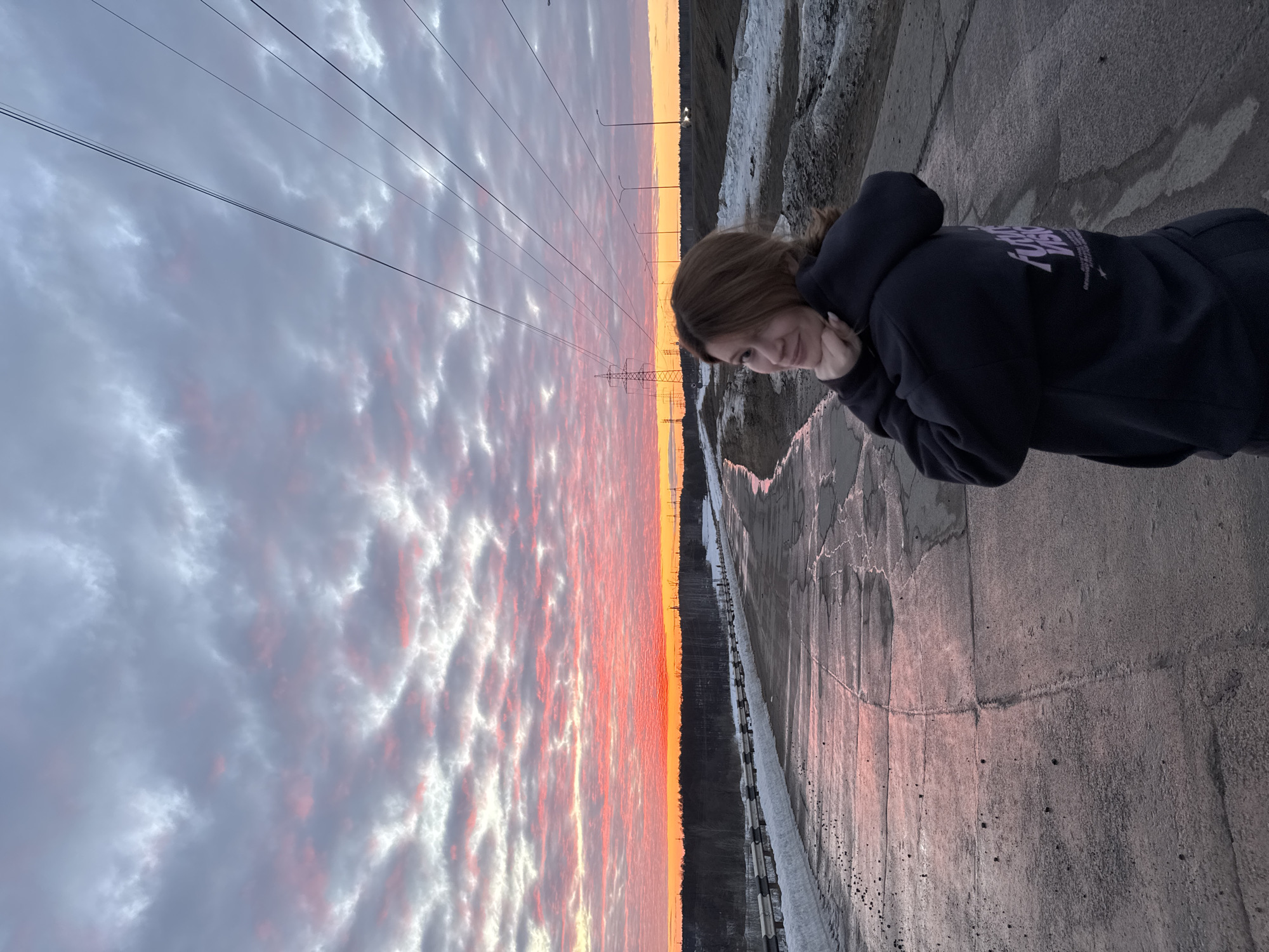 A female subject with brown hair, wearing a dark hooded sweatshirt, stands on a damp, asphalt road at sunset, likely on the outskirts of Chaikovsky, Russia. She is positioned facing away from the camera, turning her head over her right shoulder to face the observer, with a subtle smile and hands clasped beneath her chin. The road surface is uneven, showing cracks and wet patches that reflect the colorful sky. Accumulations of melting snow are present along both sides of the road. To the left, a metal guardrail runs parallel to the road, beyond which is a dense, bare forest. To the right, banks of dirt and melting snow are visible, along with multiple utility poles carrying power lines and streetlights extending into the distance. A large, lattice-type power transmission tower is visible in the mid-ground. The sky displays an extensive altocumulus cloud formation, illuminated by a sunset featuring an intense orange glow at the horizon, transitioning upwards into vibrant pink and red hues against a backdrop of gray and white clouds.