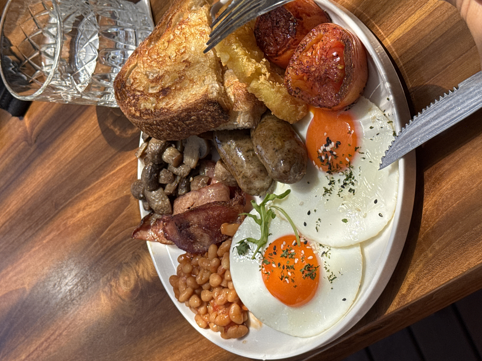 A breakfast plate, presumably in Kuta Selatan, Indonesia, sits on a wooden table. The plate contains toast, hash browns, roasted tomatoes, two sausages, two sunny-side-up eggs sprinkled with seeds and herbs, sauteed mushrooms, bacon, and baked beans. A fork is poised over the toast, and a knife rests near the eggs. A partially visible glass of water is to the left of the plate.