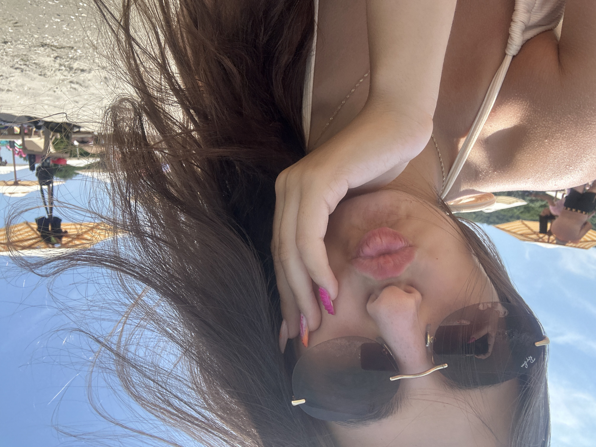 In the foreground of the image, a young woman with long, dark brown hair is taking a selfie at a beach. She is wearing large, rimless sunglasses with a gold frame and a light-colored bikini. Her hand is placed on her cheek, and her lips are pursed, as if for a kiss. Her fingernails are painted with a colorful, gradient design of pink and orange. Her hair is blowing in the wind, obscuring part of her face and the background.
The background of the image depicts a sunny beach scene. Several beach umbrellas, typical of Varna's beaches, are visible, along with beach chairs and a few people in the distance enjoying the day. The sea and the sky are not clearly visible due to the angle of the selfie and the woman's hair. The overall impression is a casual, summery snapshot taken on a bright day.