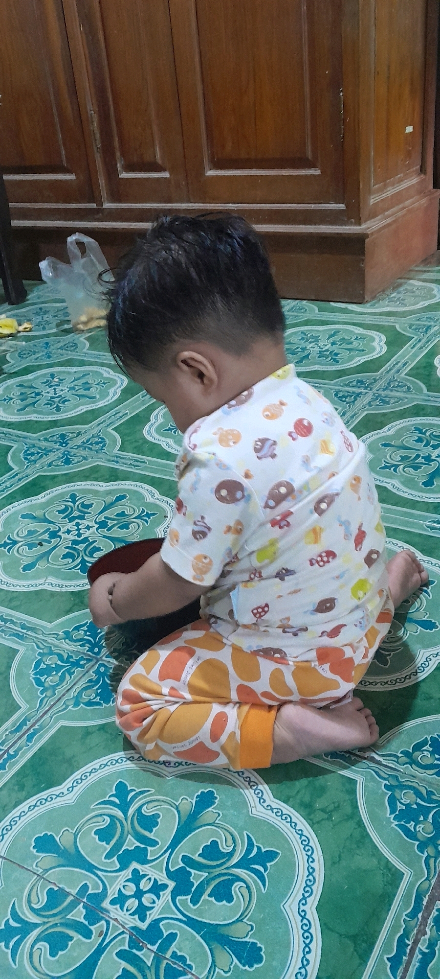 In Sambong, Indonesia, a young child, viewed from behind, is sitting on a green tiled floor. The child is wearing a white t-shirt with a repeating pattern of cartoon fish and orange pants with a pattern of ovals. The child is leaning forward, looking down at a dark bowl in their hands. The floor is decorated with a repeating pattern of ornate, light-green and dark-green floral designs. In the background, a wooden cabinet with paneled doors is visible. A white plastic bag lies on the floor near the cabinet.