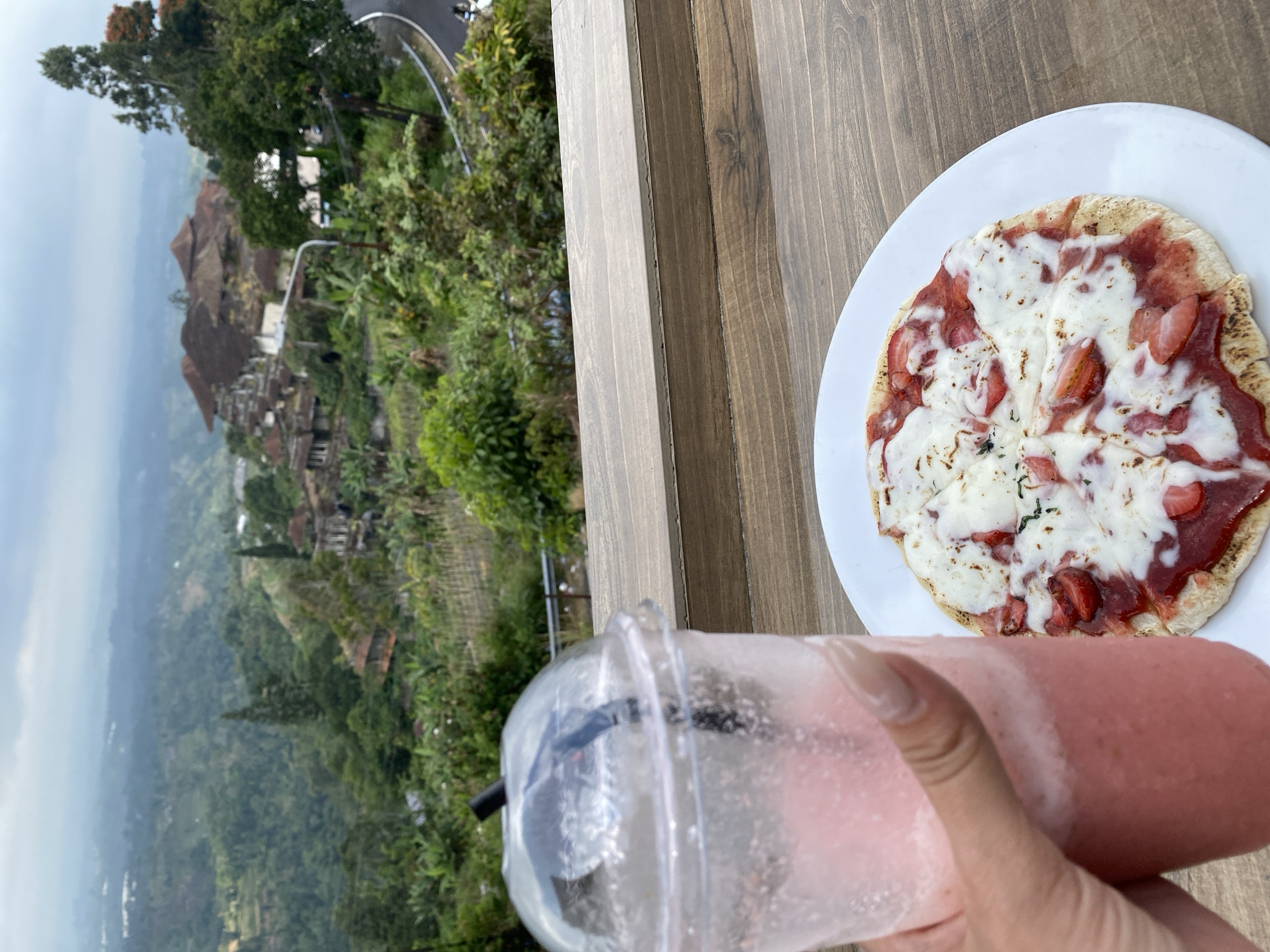 A hand holds a clear plastic cup filled with a pink, blended beverage, likely a smoothie, with a black straw inserted. The cup is positioned on a wooden surface in the foreground. To the right, a white plate holds a small pizza topped with melted white cheese and sliced strawberries. The pizza is also on the wooden surface. In the background, a lush green landscape with rolling hills and scattered buildings is visible under an overcast sky. The scene appears to be at a scenic overlook, potentially in or near Baturiti, Indonesia, given the tropical vegetation and architectural style of the distant structures.