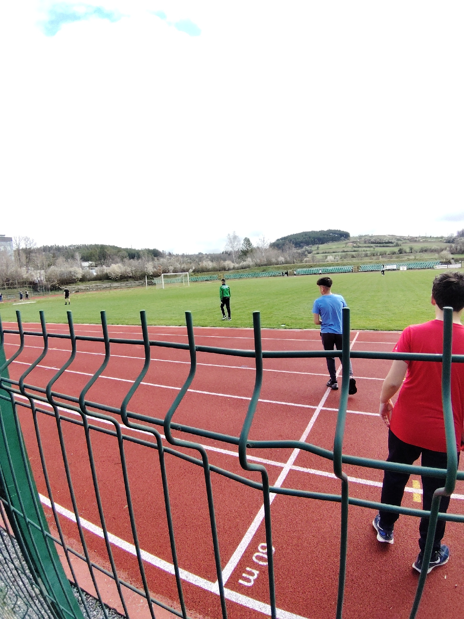 The image shows a sports field with a running track in Panagyurishte, Bulgaria. A green fence runs along the foreground of the track. On the track, two young men are walking. One wears a red t-shirt and black pants, while the other wears a blue t-shirt and dark pants. Another person in a green uniform stands on the grass field beyond the track. A soccer goal is visible on the grass field, and several people are scattered in the background. The landscape features trees and a distant hill under a cloudy sky.