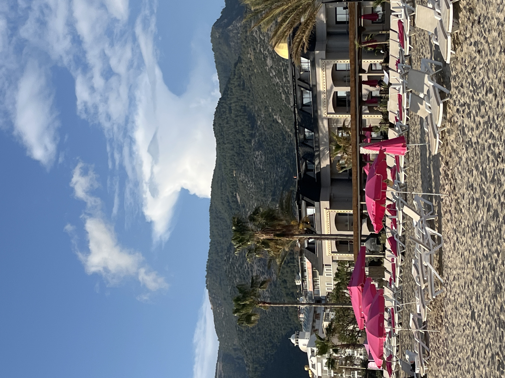 The image shows a beach scene in Alanya, Turkey. A large mountain rises in the background next to a multi-story building. Palm trees are interspersed between the mountain and the building. The foreground is filled with beach umbrellas and white beach chairs. The sand appears light in color. The sky is blue with scattered white clouds.