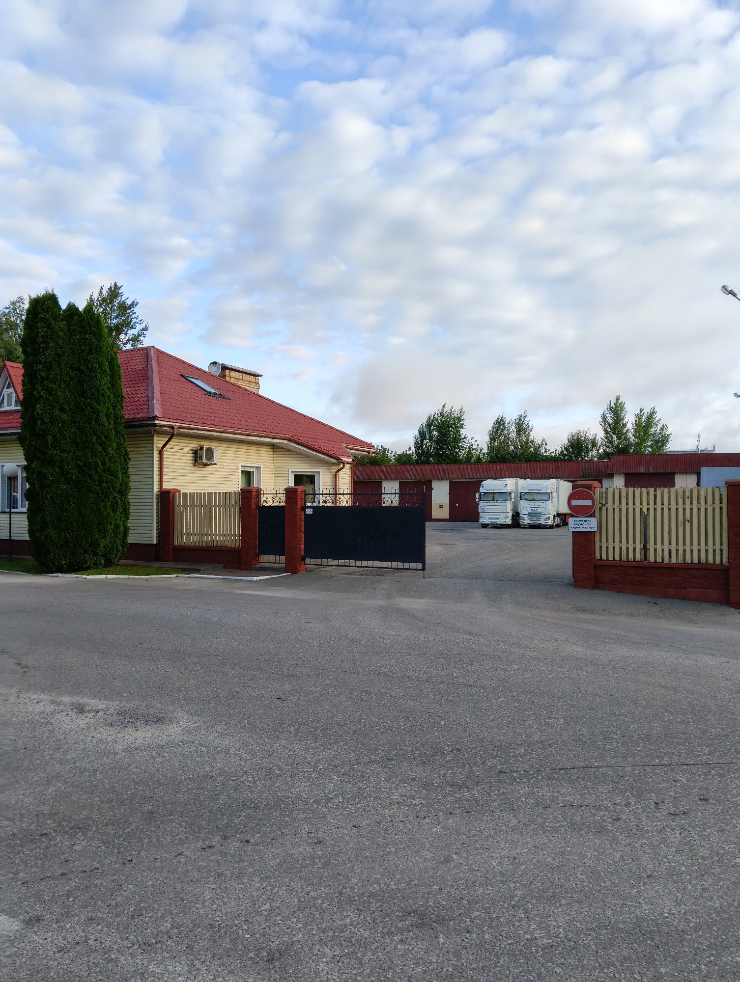 The image depicts a scene in the city of Hrodna, Belarus, featuring a residential building with a red roof and light yellow siding, adjacent to a tall evergreen tree. A fence with brick pillars and a wooden section separates the property from a paved area. Beyond this fence, a gate leads into a lot containing several large warehouse-like structures with red trim. Two white semi-trailer trucks are parked in front of these buildings. A circular red and white "no entry" sign is visible on a wooden fence to the right of the trucks. The sky above is cloudy. The overall environment suggests a mixed-use area with residential and commercial or industrial elements.