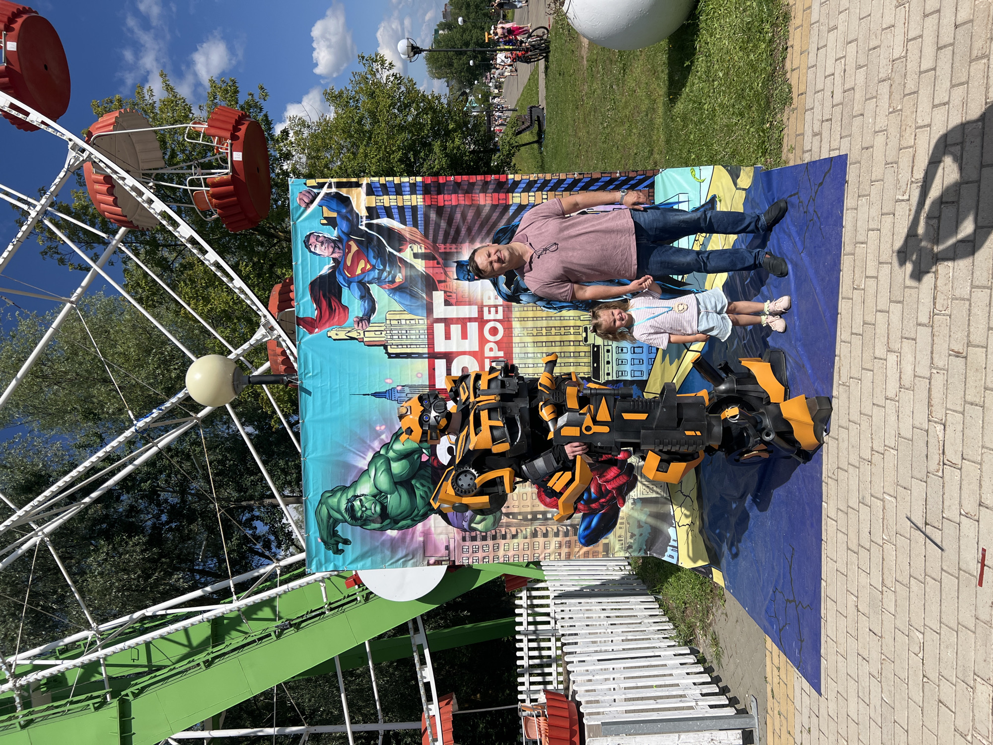 This image depicts an outdoor scene, likely in a park or amusement area, with a ferris wheel visible in the background. In the foreground, a person in a Bumblebee costume from Transformers stands next to a backdrop featuring comic book characters such as the Hulk and Superman. A man and a young girl are posing for a photo in front of this backdrop, holding hands. The ground is paved with brick. The location is identified as Cheboksary, Russia.