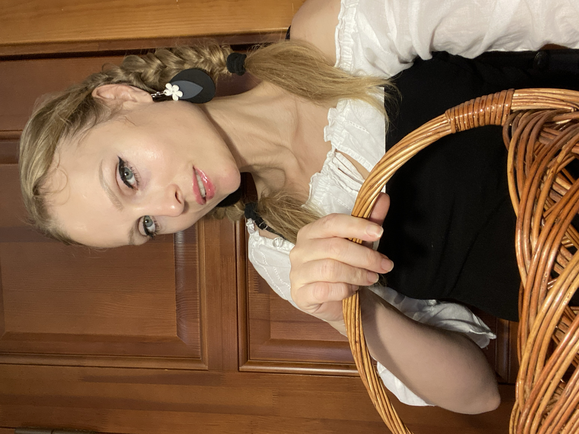 The image shows a medium shot of a young, fair-skinned woman with blonde hair styled in two pigtails. She is wearing a white, off-the-shoulder blouse with ruffled trim and a black dress or top underneath. She has on large, dark teardrop-shaped earrings with a white flower accent. The woman is holding a large, woven basket in front of her. Her left hand is visible, resting on the rim of the basket. She has noticeable makeup, including winged eyeliner and glossy pink lipstick. The background is a wooden surface, likely a door or wall paneling, with vertical lines and a visible hinge on the left. The lighting suggests an indoor setting. There is no indication of the city of Lida, Belarus, in the image itself.