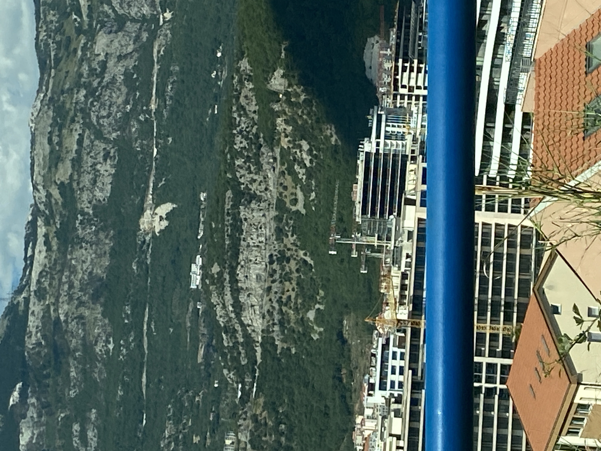 This is a vertical, medium-shot, eye-level photograph taken from a distance, capturing a panoramic view of Budva, Montenegro. In the foreground, a bright blue horizontal railing runs across the lower portion of the image. Behind the railing, a section of a building with a terracotta tiled roof and cream-colored walls is visible, with some greenery in front of it.
Further back, a line of modern buildings forms the urban landscape. These structures vary in height and architectural style, with some featuring dark-tinted windows and balconies. Construction cranes are interspersed among the buildings, indicating ongoing development. The city stretches along the base of a large, densely forested mountain that dominates the background. The mountain's rugged terrain is characterized by rocky outcrops and patches of green vegetation, with a few scattered buildings visible on its slopes. The sky above is a mixture of blue and white clouds. The lighting suggests it is daytime, possibly late afternoon, as evidenced by the shadows cast on the mountain.