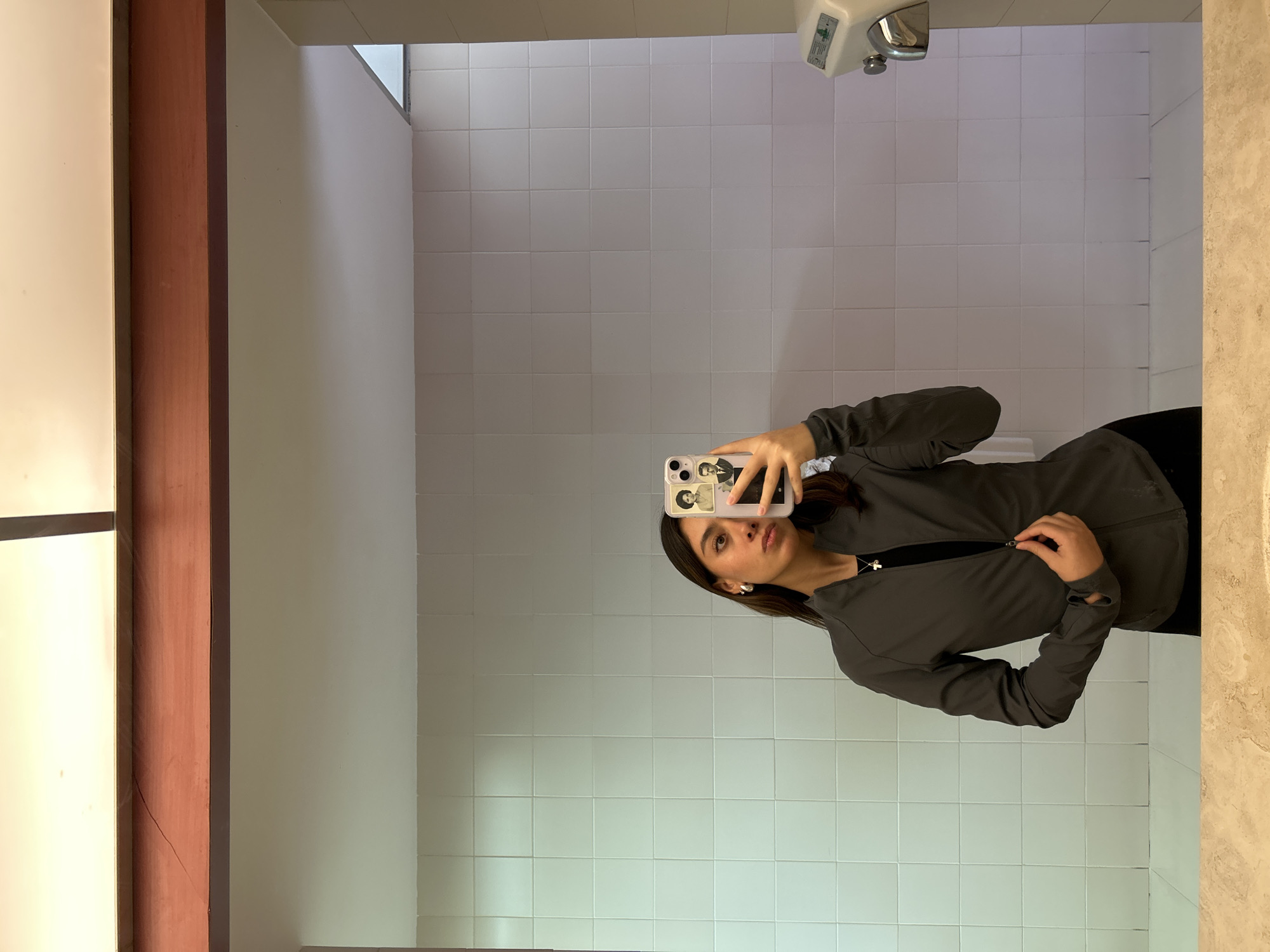 A person is taking a mirror selfie in a bathroom with white tiled walls. The person is holding a phone with a case that appears to have images of historical figures on it. They are wearing a dark-colored zip-up jacket and a necklace with a small cross pendant. The bathroom has a hand dryer mounted on the wall. A light-colored surface, possibly a countertop, is visible on the right side of the frame. The bathroom also has a light-colored wall and a wooden door frame to the left.