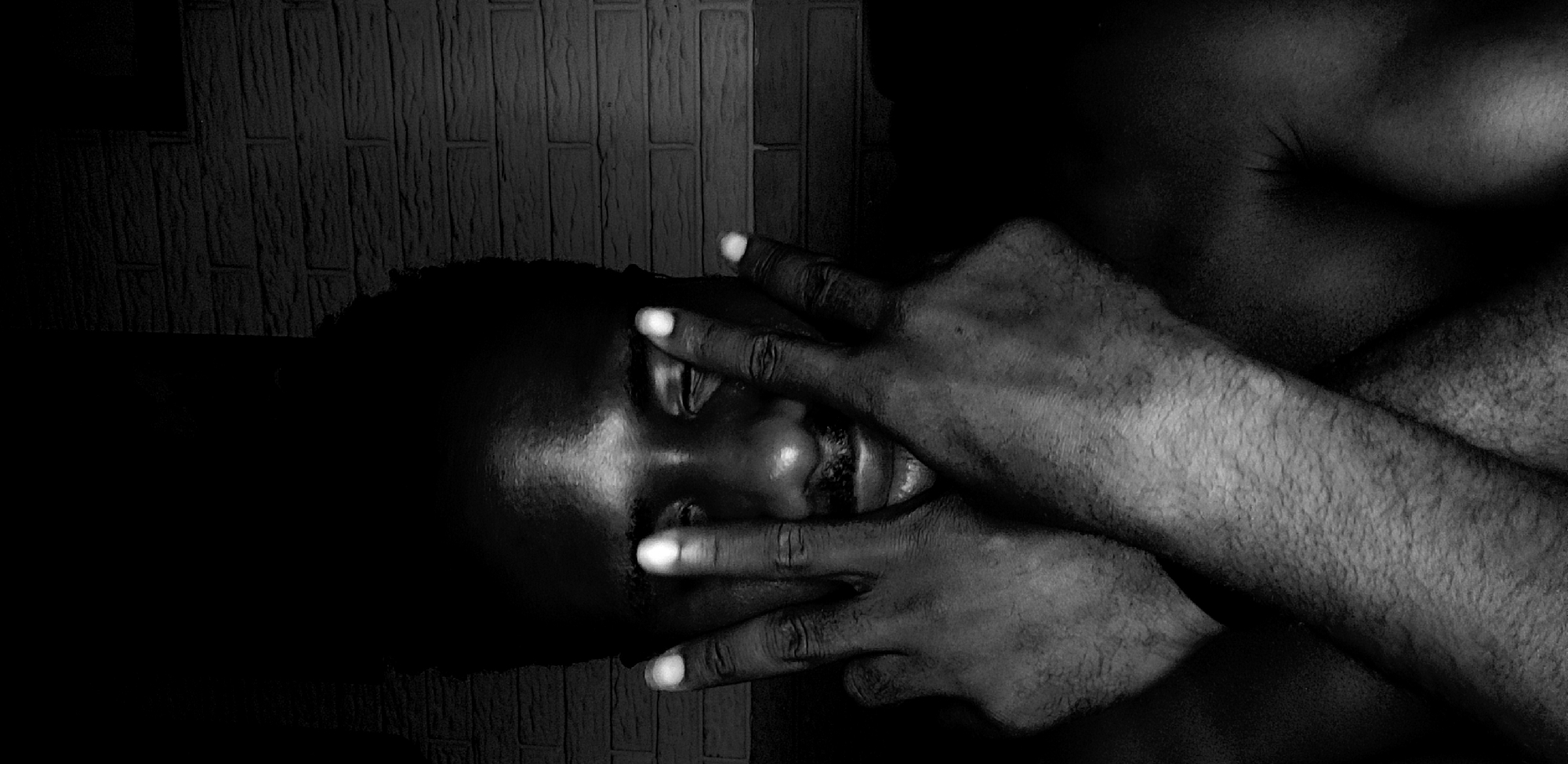 A striking black and white, low-key portrait features a person, likely male, positioned horizontally and partially obscuring their face with their hands. The subject's dark skin tone is rendered with strong contrasts, highlighting features such as the forehead, nose, and slightly parted lips, which reveal a hint of teeth. A faint mustache is visible.
Both hands are placed over the face, with fingers spread across the eyes and cheeks. The fingertips are notably brighter, almost luminous, creating a distinct visual focus against the darker skin. The hands show texture and subtle hair. Parts of the person's upper torso and shoulder are visible, blending into the deep shadows.
The background is a dark, vertically textured surface, possibly a wall, contributing to the intimate and somewhat somber atmosphere. The lighting is dramatic, characterized by deep shadows and sparse highlights, which accentuate the contours of the subject and their hands.
The activity depicted is the person covering their face, which conveys a sense of introspection, shyness, vulnerability, or perhaps weariness. The overall mood is contemplative and melancholic. No text is visible in the image. The setting is in Narayi, Nigeria.