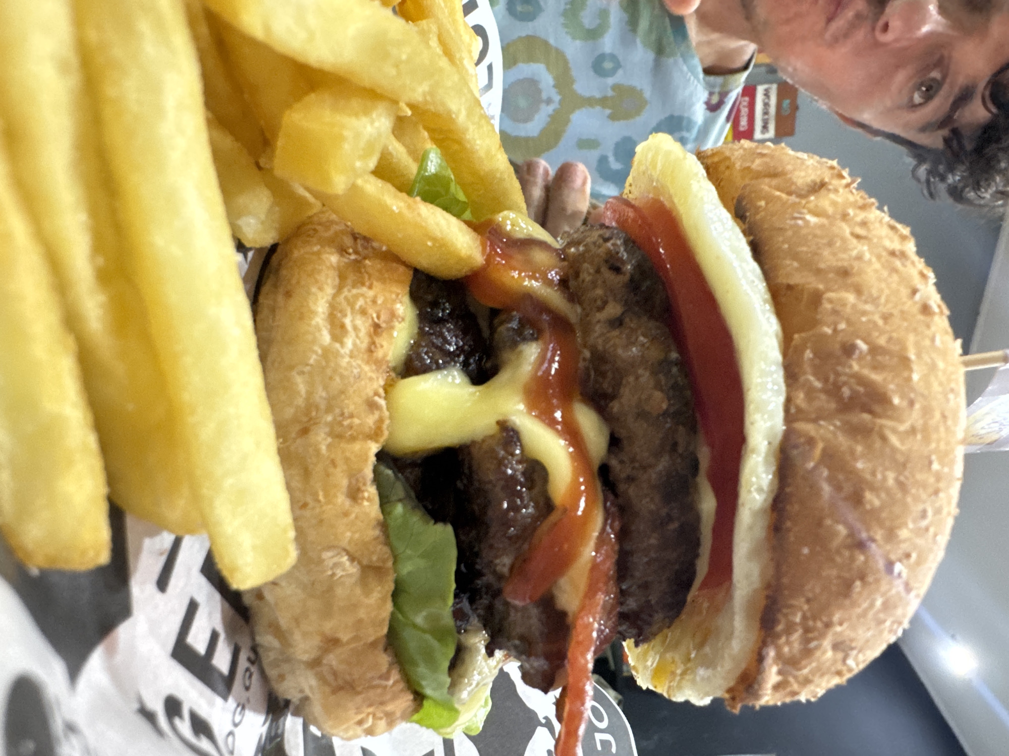 A close-up shot shows a hamburger and French fries, likely taken in Kuta, Indonesia. The hamburger is in focus, revealing visible lettuce, a meat patty, melted cheese, and ketchup. The fries are stacked beside the hamburger, seemingly overflowing from a container underneath. The background features a blurred individual, partially in focus. The food sits on a printed wrapper. The environment around the food is a mix of colors and textures, suggesting an indoor setting with natural lighting.