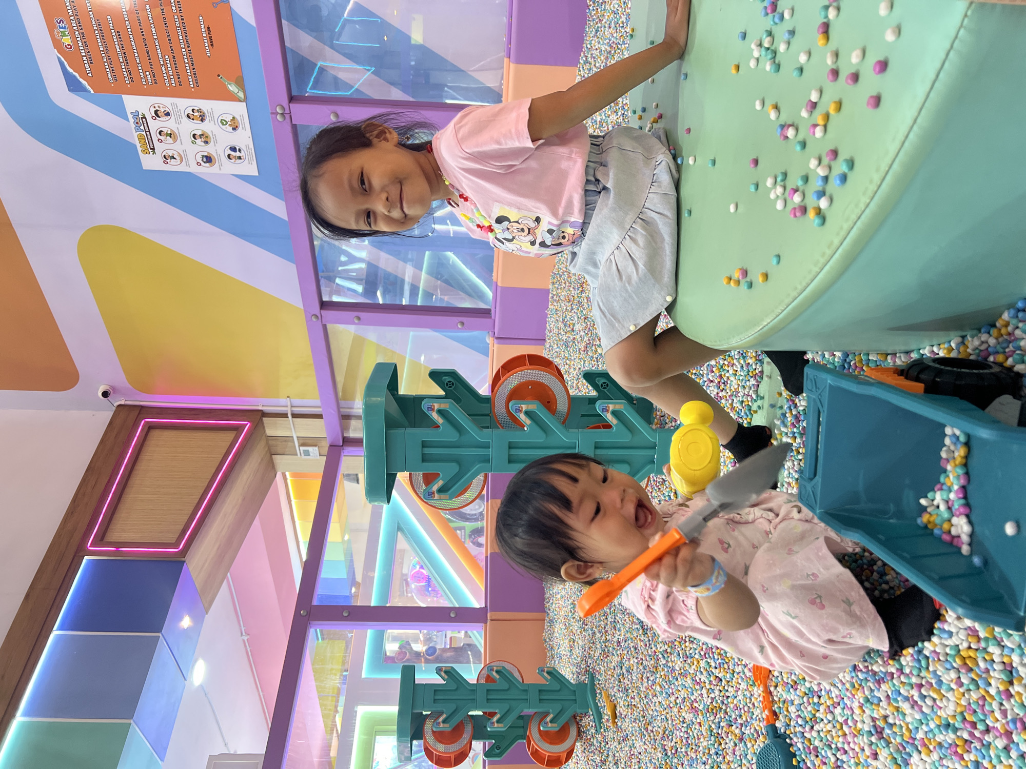 This photograph captures a lively indoor playground setting, likely located in Kecamatan Pati, Indonesia. The scene is dominated by a ball pit filled with countless colorful, small balls in shades of pink, blue, yellow, and white. In the foreground, a young child, appearing to be a toddler, is kneeling and actively engaged in play. The child is wearing a pink dress with a cherry pattern and black socks. They are holding a bright orange shovel, attempting to scoop balls into a teal toy dump truck. Adjacent to the dump truck is a yellow toy watering can.
To the right, an older child, a girl, is sitting on a rounded, mint-green structure that serves as seating within the play area. She is wearing a pink t-shirt with a Mickey Mouse graphic and a light blue, ruffled skirt. She is smiling and looking towards the camera. Her outfit is accessorized with a colorful beaded necklace.
The background reveals a vibrant play structure with towering, teal, tree-like elements that have orange wheels on their sides. Beyond this, the environment suggests a multi-level play area with transparent purple and pink barriers, and colorful, geometric wall decorations illuminated by neon lights. A sign is visible on the wall behind the older child, containing text and illustrations related to playground rules. The overall impression is one of a bright, energetic, and child-friendly entertainment space.
