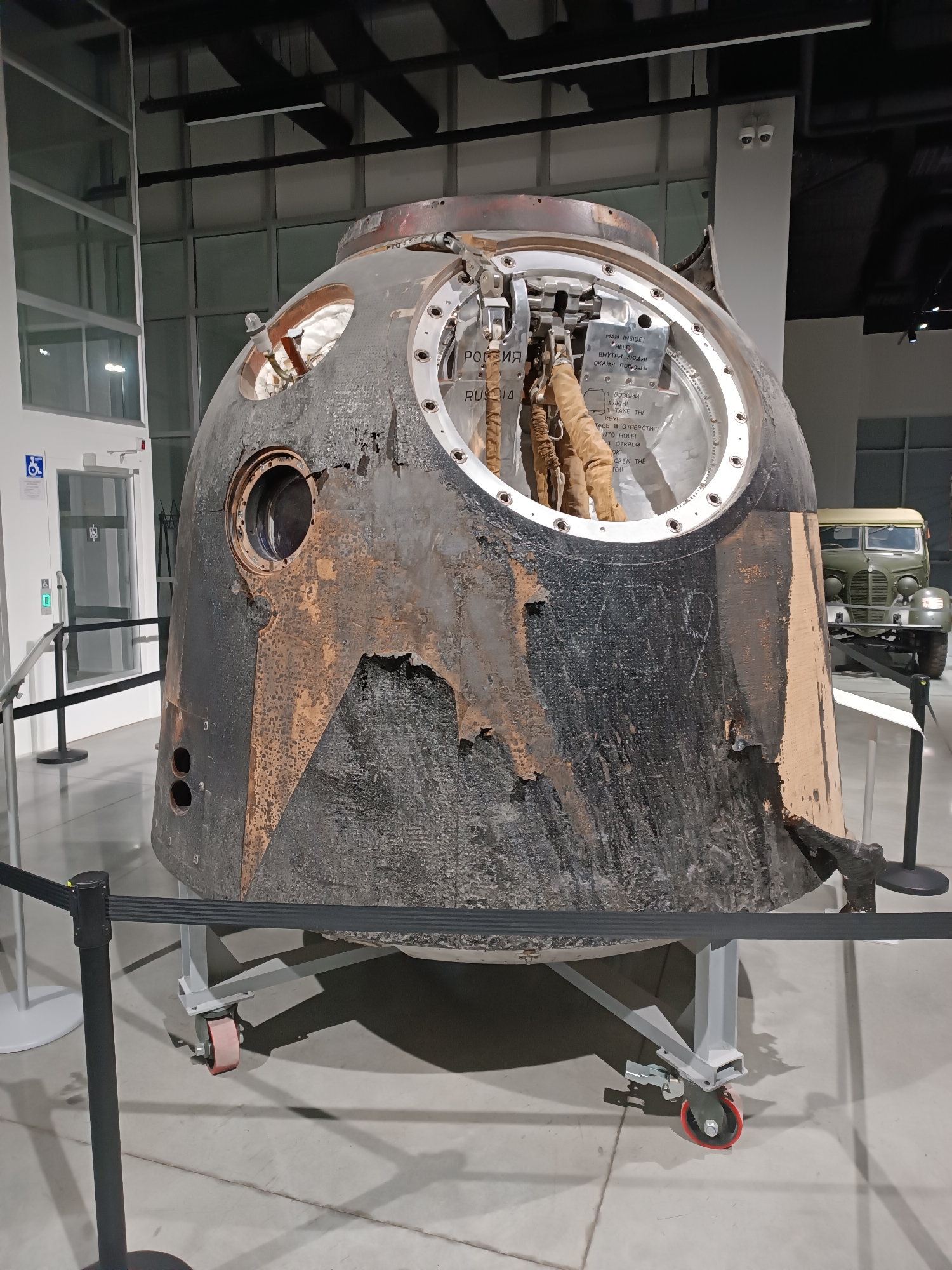 The image shows a Soviet-era spacecraft capsule, identified by the word "РОССИЯ" (RUSSIA) inscribed on its exterior, displayed on a wheeled stand. The capsule appears to be scorched and weathered, with sections of its outer hull missing or damaged, revealing internal components. Inside the open hatch, tan-colored straps and cables are visible. The spacecraft is positioned within a museum setting, indicated by the polished concrete floor, modern lighting fixtures, and a visible wheelchair-accessible symbol on a nearby door. In the background, a vintage military truck is also on display. The location is identified as the Museum of Military Technology in Verkhnyaya Pyshma, Russia.