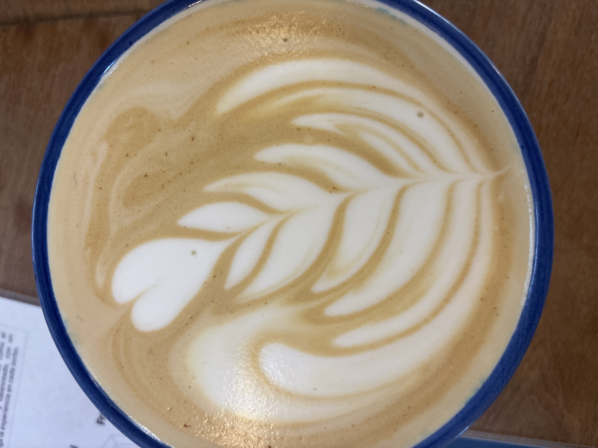 A close-up, overhead shot depicts a cappuccino or latte with latte art resembling a fern or leaf pattern in a blue-rimmed cup. The coffee sits on a wooden surface. A partially visible document with text in Spanish is present in the lower left corner. The setting appears to be a cafe or restaurant. The location in the city of Buenos Aires, Argentina, is unknown.