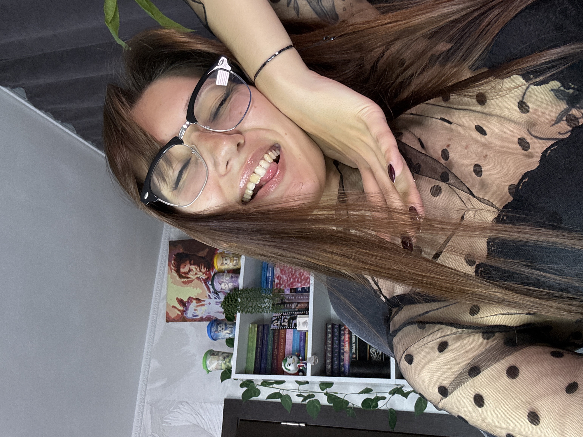 The image shows a close-up, selfie-style shot of a young woman indoors. She is wearing glasses and a sheer black top with polka dots over a black bra. Her right hand is resting on her cheek, and her tongue is sticking out slightly. Behind her, a white bookshelf is visible, filled with books and decorative items, including what appears to be a painting of Iron Man. A potted plant with green leaves is also visible on the right side of the image. The lighting suggests an indoor environment, possibly a room in a residential apartment. The overall aesthetic is casual and playful. There is no information in the image to indicate the specific location in Novokuznetsk, Russia.