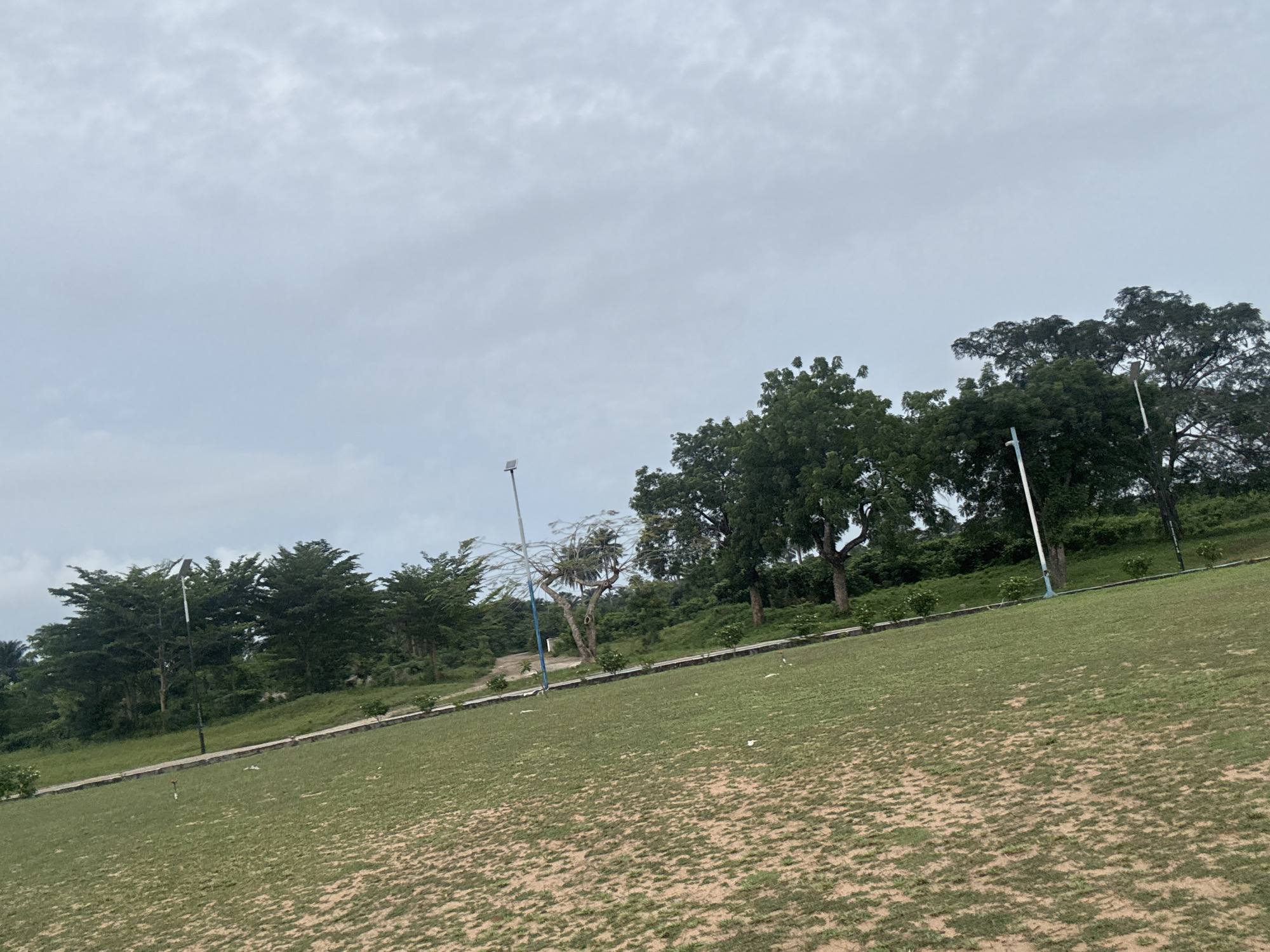 The image shows a park in Ihiagwa, Nigeria, on an overcast day. The foreground is dominated by a grassy field interspersed with patches of exposed soil. A pathway runs across the slope. Trees of varying sizes and types are visible along the horizon, with several lampposts positioned along the path and within the trees. The sky is uniformly cloudy. There are no people visible.