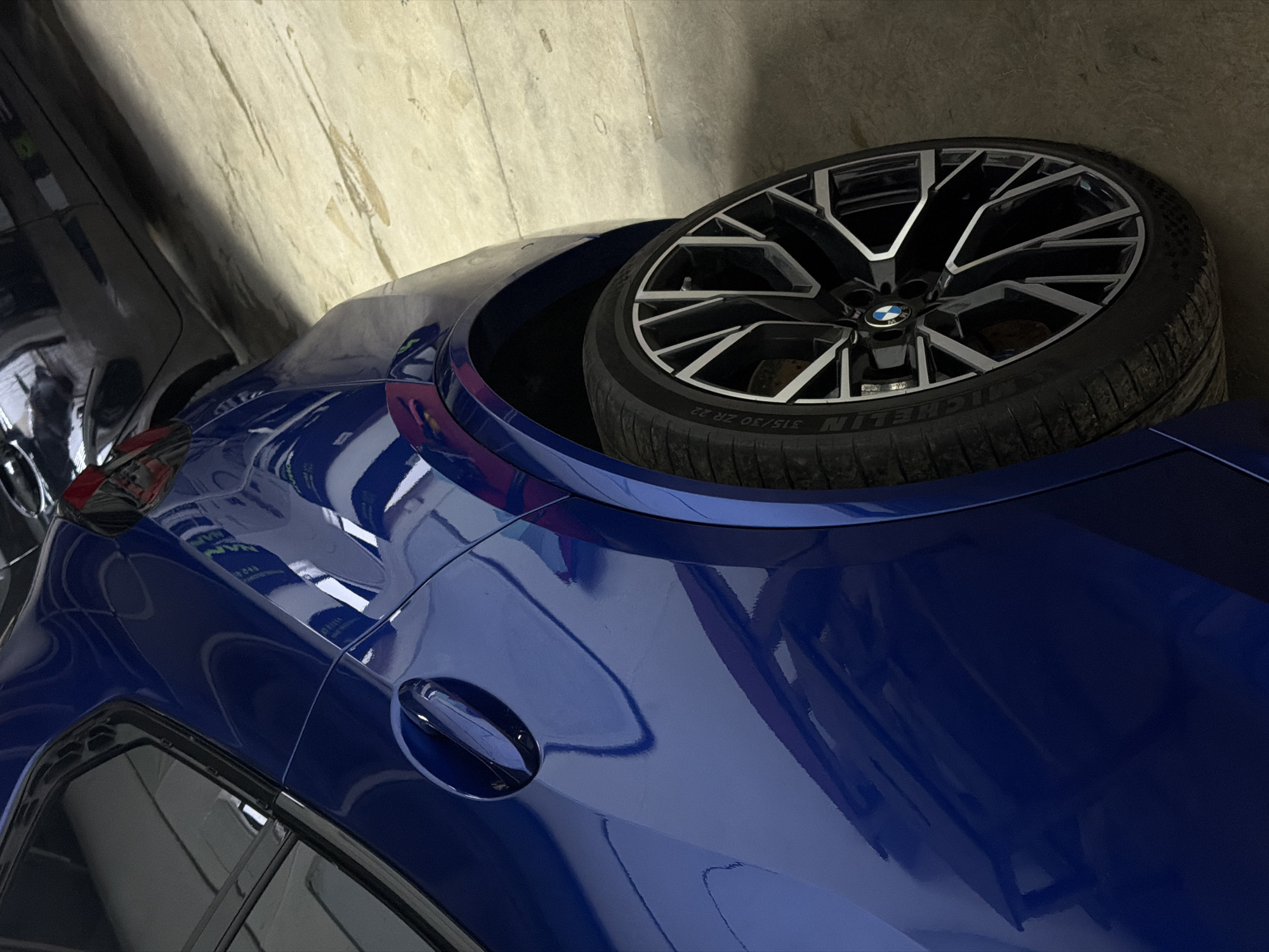 The image shows a close-up of the rear of a blue BMW car. A tire with a rim is sitting on the rear fender. The car is parked in what appears to be an indoor garage or parking structure with a beige wall visible in the background. It is not possible to determine if the location is in Krasnoyarsk, Russia, or the actions, interactions, or other objects from the image.