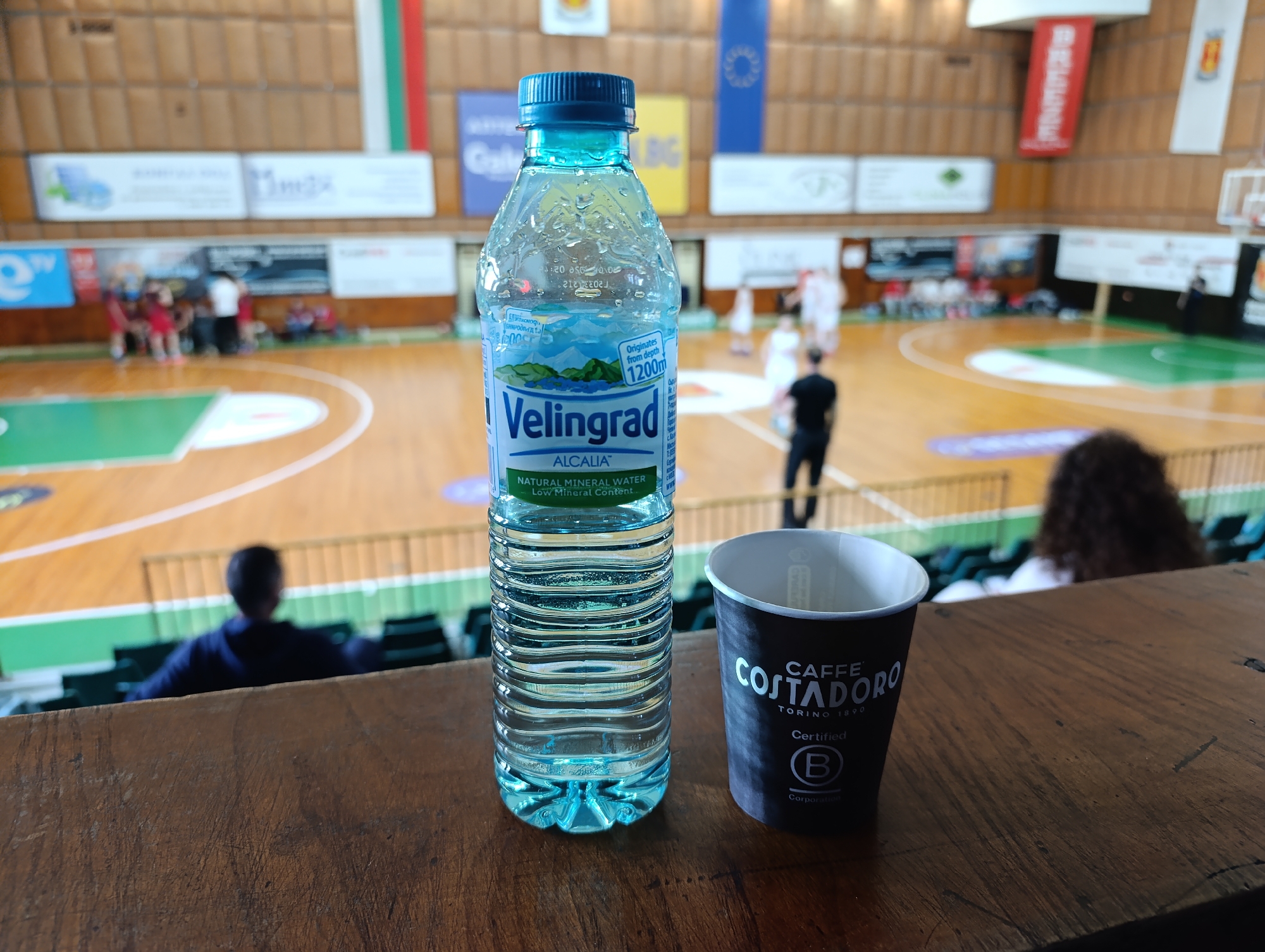 The image presents a scene at a basketball court in Haskovo, Bulgaria. In the foreground, a bottle of Velingrad water and a Caffe Costadoro coffee cup rest on a wooden surface. The basketball court is visible in the background with players and spectators present. Bulgarian and European flags are displayed near the ceiling.