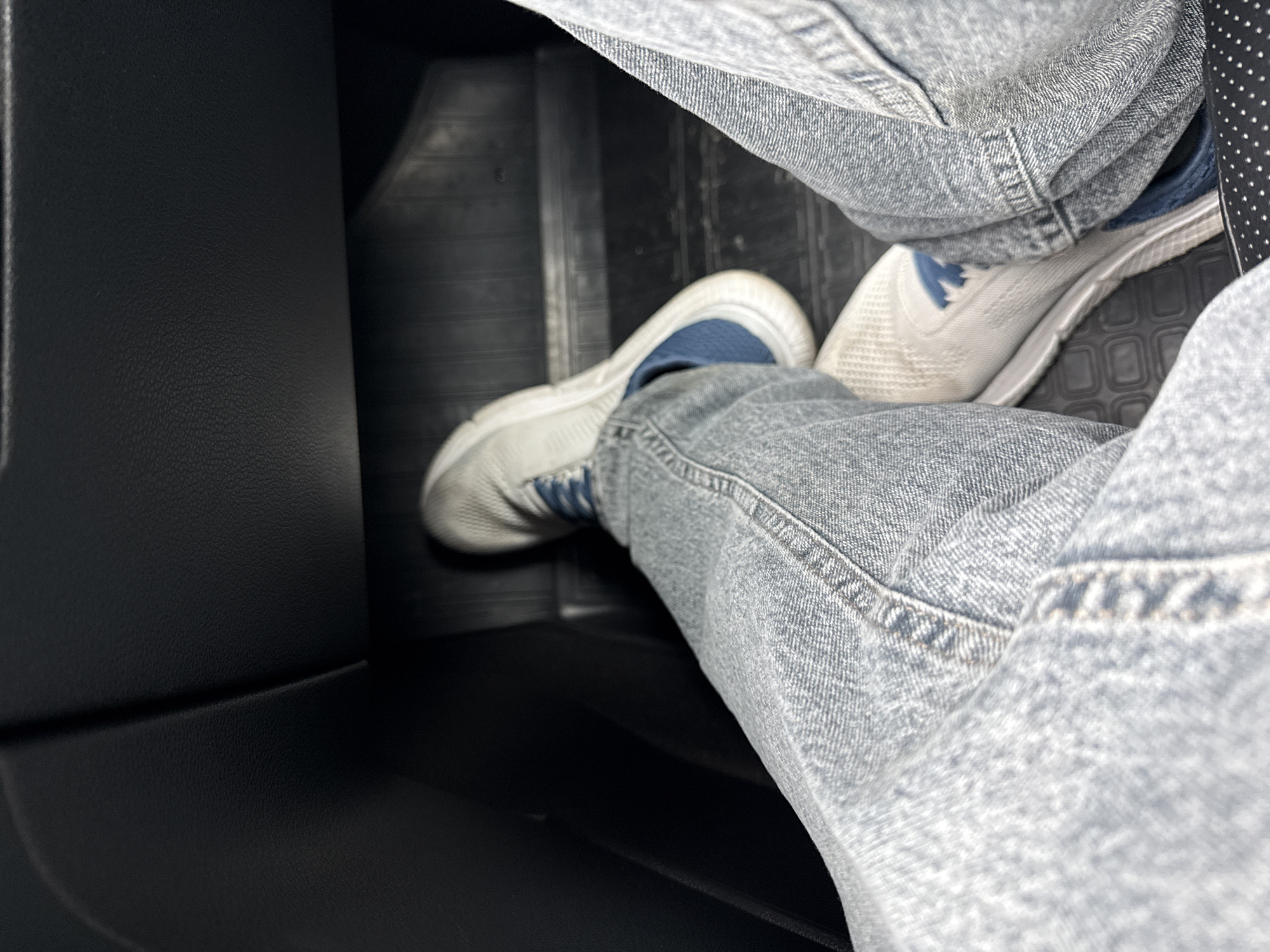 The image shows a person seated in the interior of a vehicle. The person is wearing light-wash denim jeans and white athletic shoes with blue accents. The flooring of the vehicle is black. A portion of the black interior, including the seat, is visible in the left side of the frame. The location is not discernible from the image and cannot be confirmed as Sochi, Russia.